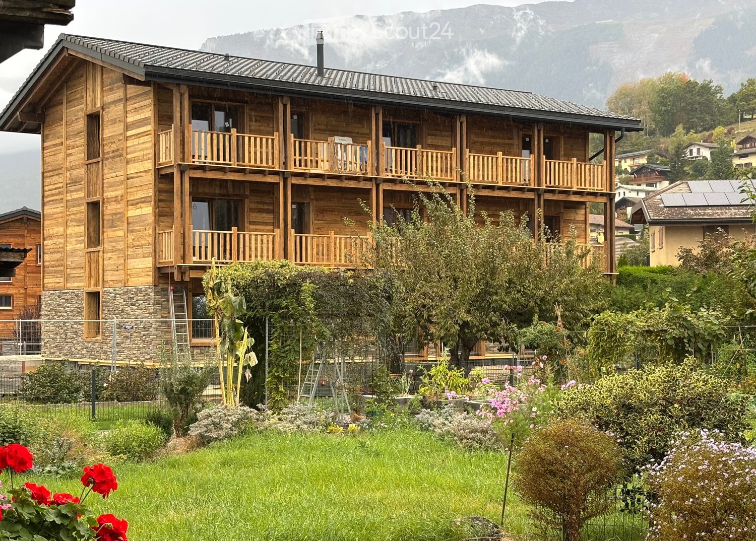 A multi-story wooden house with balconies, surrounded by a garden with flowers and trees, with mountains in the background.