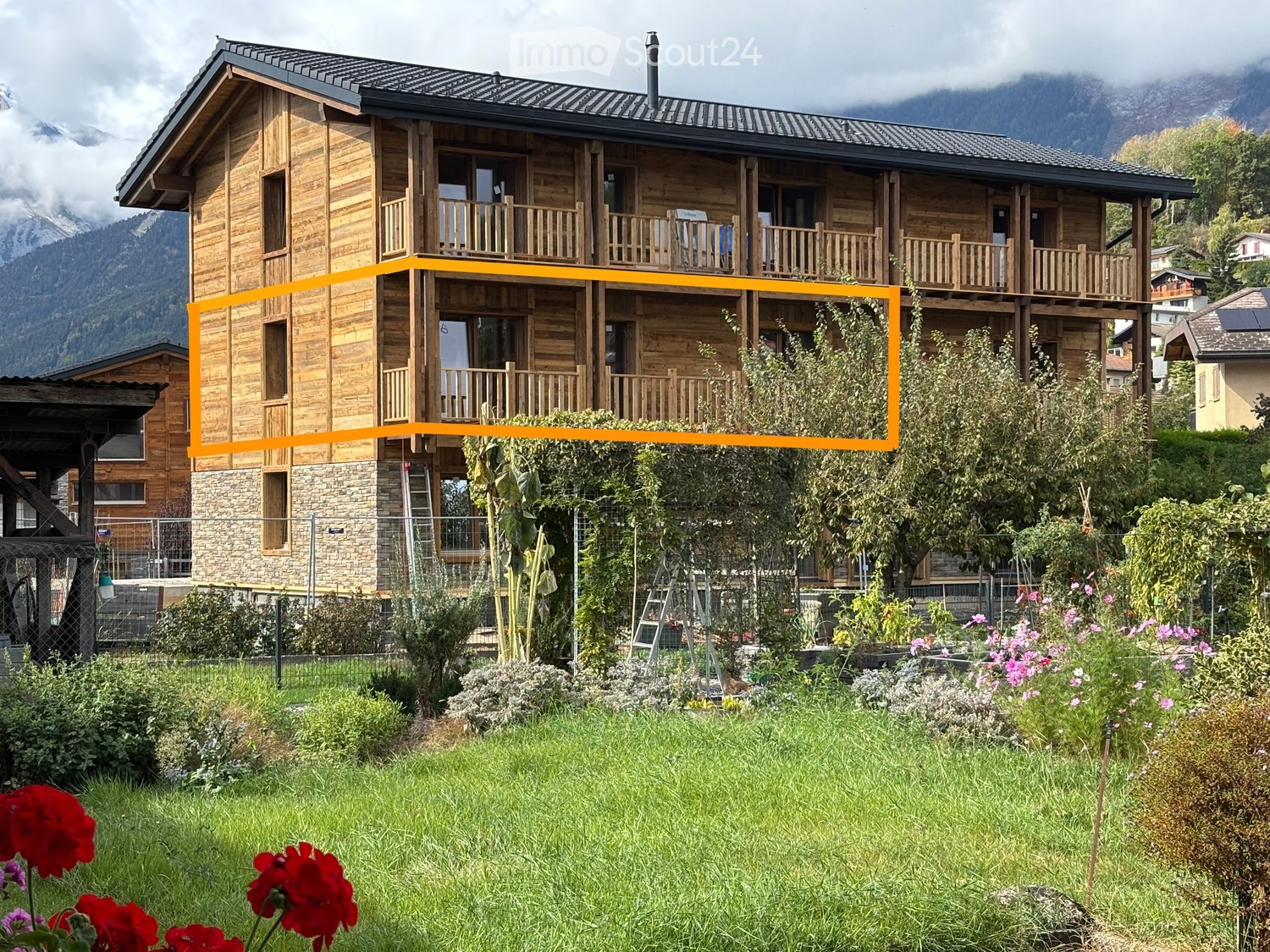 A two-story wooden house with balconies, surrounded by a garden with flowers and trees, set against a mountainous backdrop.