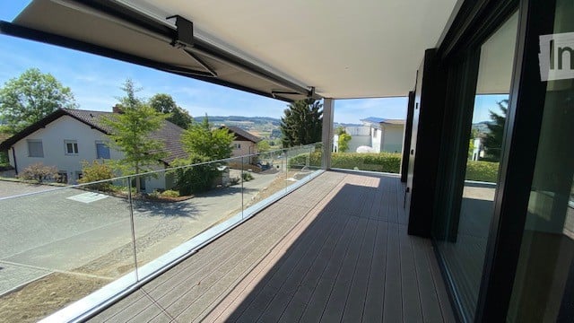 Modern balcony with wooden decking, glass railing, and scenic view of surrounding houses and landscape.