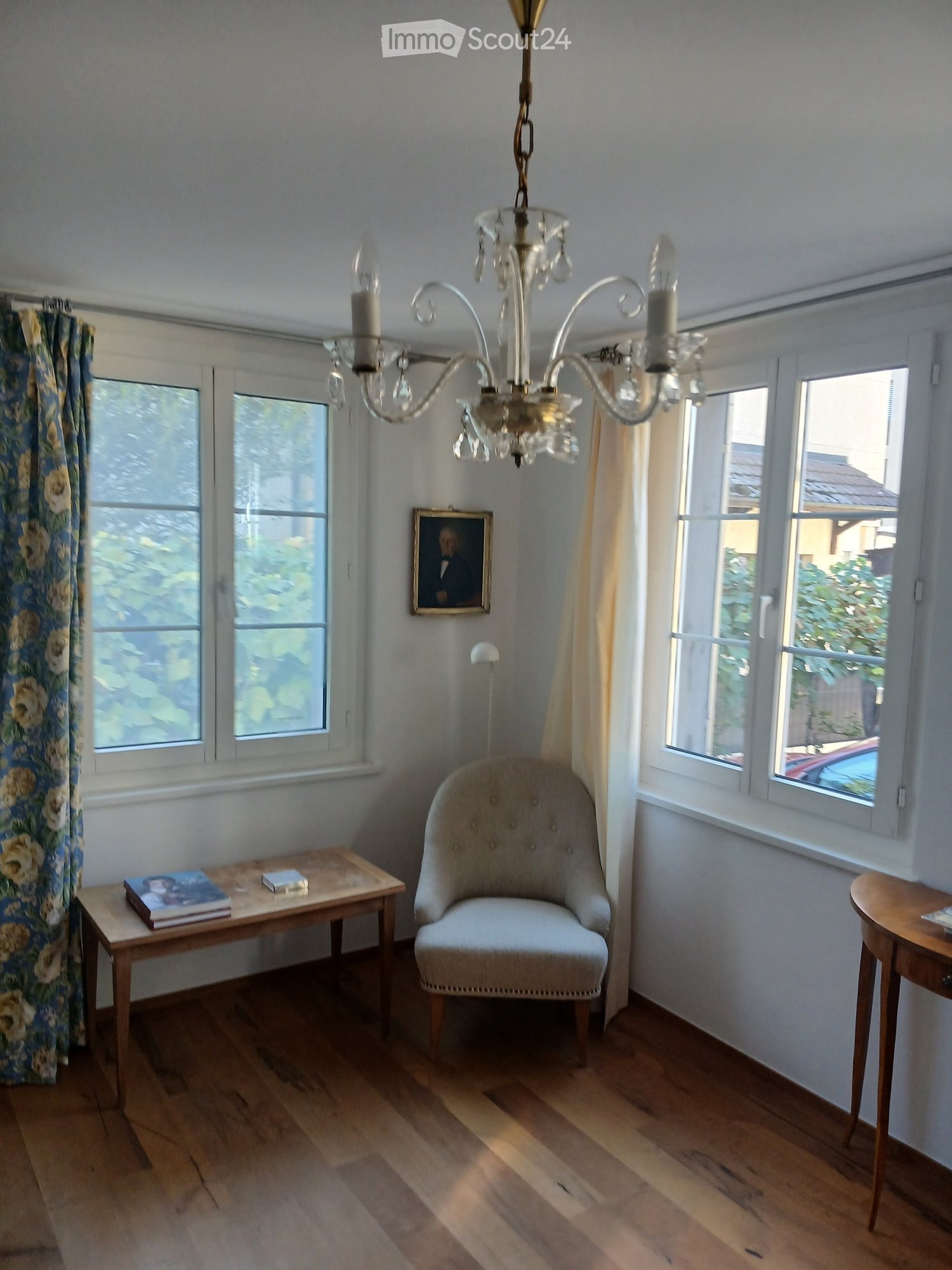 Bright room with wooden flooring, large windows, a chandelier, a single armchair, a small table with books, and a framed picture on the wall.