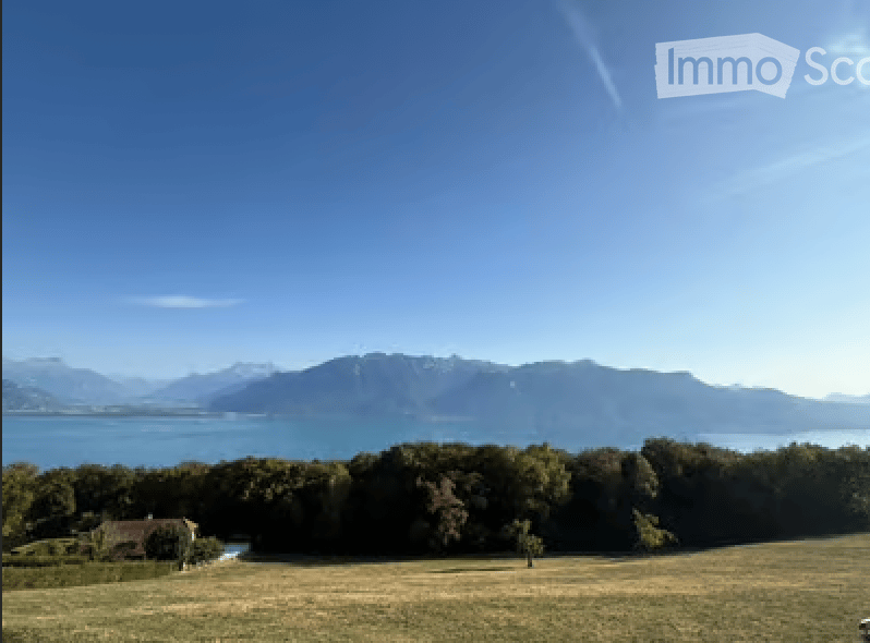 Open field with trees, a small structure, and a distant lake and mountains under a clear blue sky.
