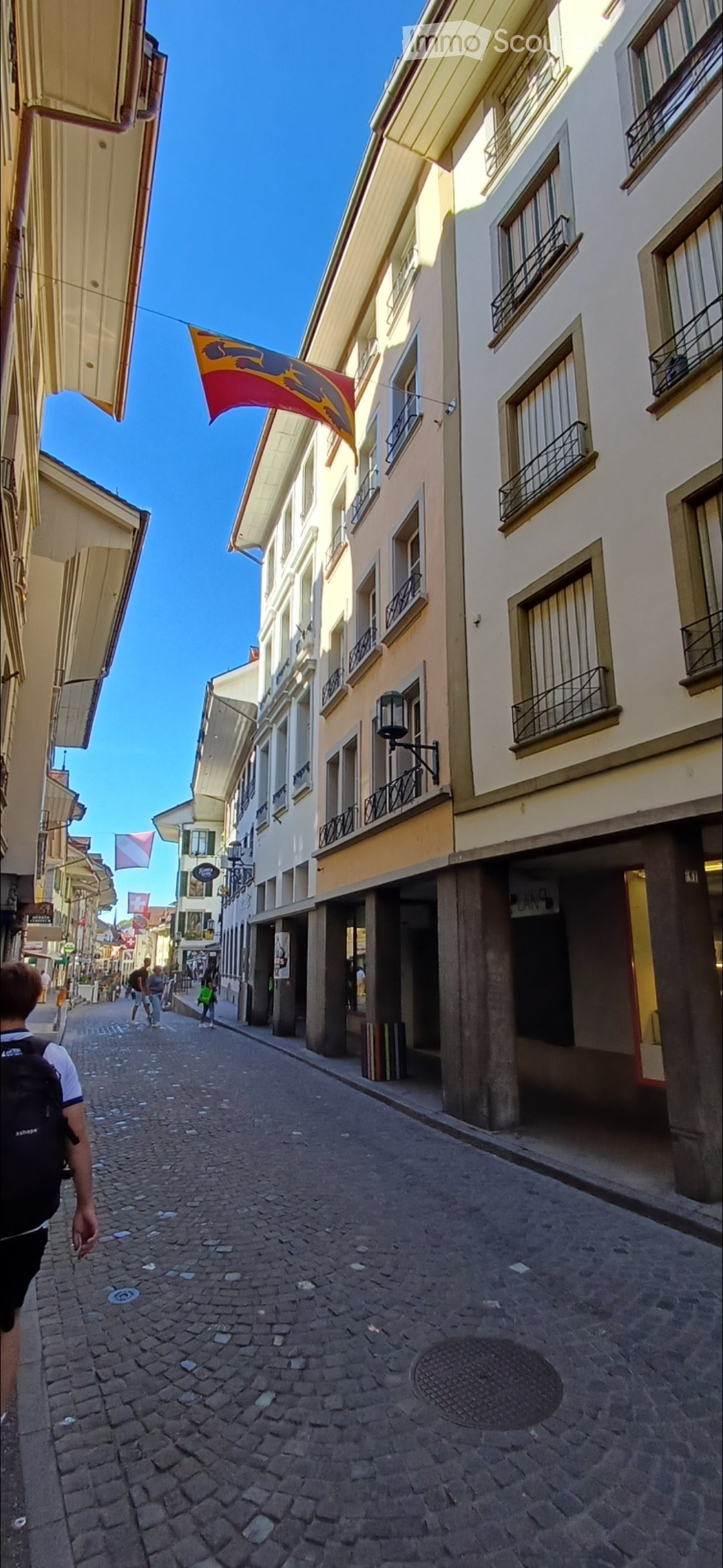 A narrow cobblestone street lined with multi-story buildings featuring balconies and awnings. A flag is visible above one of the buildings.