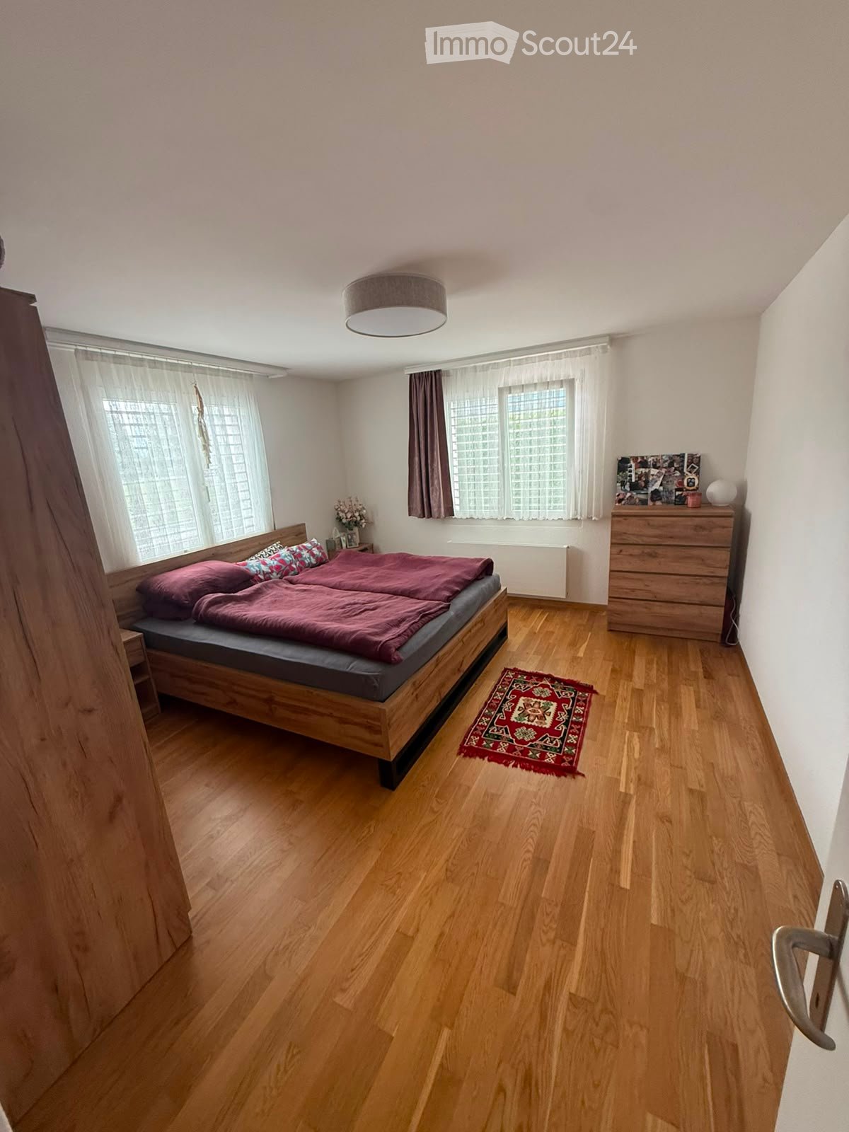 A well-lit bedroom with wooden flooring, a double bed with red bedding, a nightstand, a dresser, and a small rug near the bed.