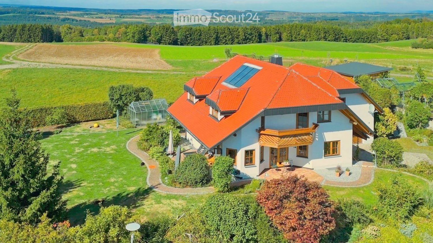 A modern two-story house with a red-tiled roof, white walls, and large windows, surrounded by a well-maintained garden and open fields.