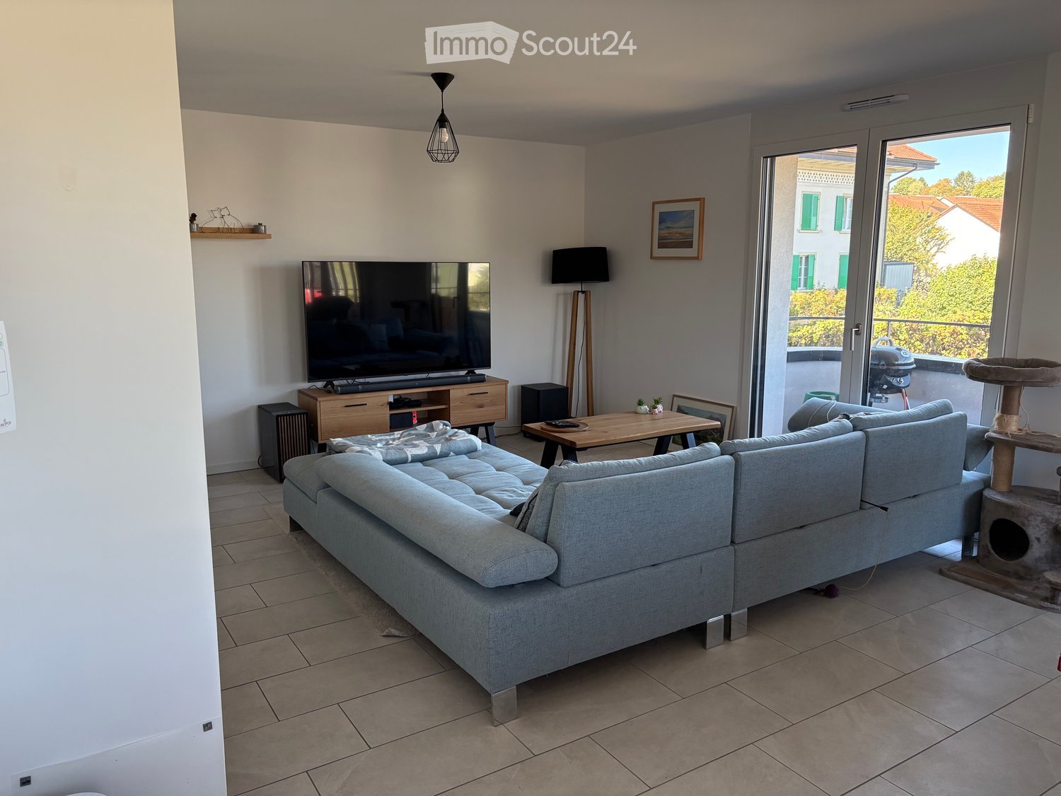 Modern living room with a grey sectional sofa, coffee table, flat-screen TV on a wooden stand, floor lamp, cat tree, and a view through large glass doors to a balcony.