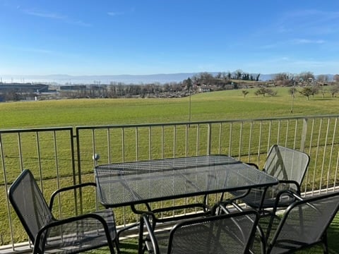 Balcony with a metal railing, overlooking a green field and distant hills. A glass-top table and four metal chairs are placed on the balcony.
