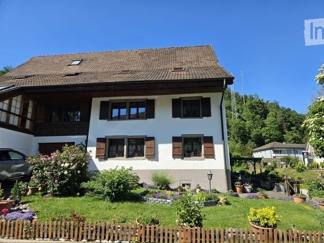 A two-story house with a brown tiled roof, white walls, and multiple windows with wooden shutters. The house is surrounded by a well-maintained garden with various plants and flowers, and a wooden fence borders the property.