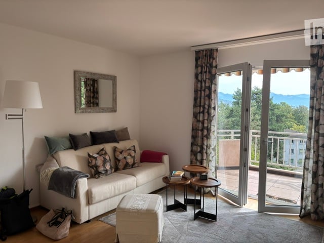 Bright living room with large windows and sliding glass doors leading to a balcony with a scenic mountain view. Features a white sofa with multiple throw pillows, an ottoman, a coffee table, side tables, and a floor lamp.