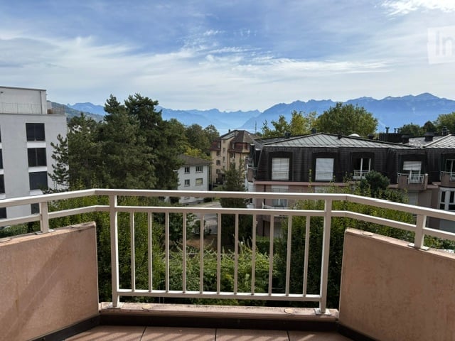 Balcony with white metal railing, overlooking a cityscape with trees and distant mountains.