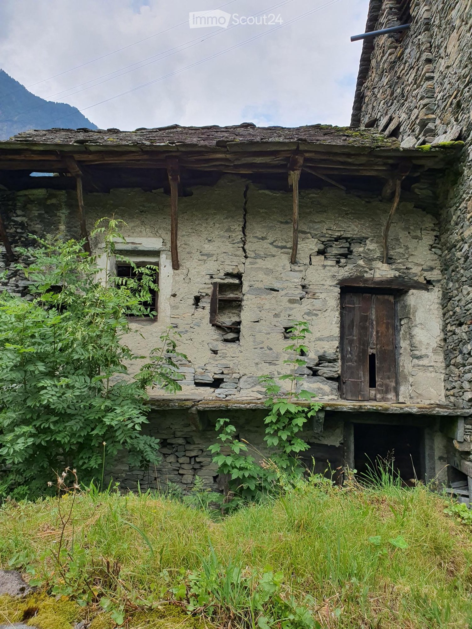 A rustic stone house with a wooden door, small window, and overgrown vegetation around it, situated in a grassy area with a mountainous backdrop.