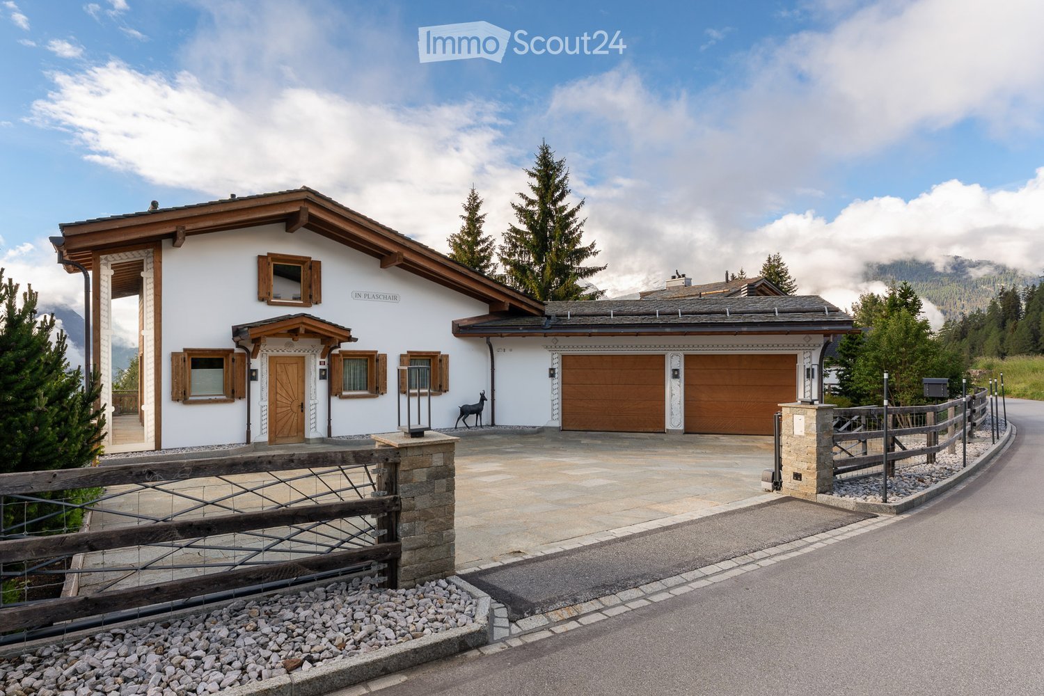A two-story house with a white exterior, wooden garage doors, and a stone driveway. The house features a traditional alpine design with wooden shutters and a sloped roof. There is a decorative wooden fence and a small statue near the entrance.