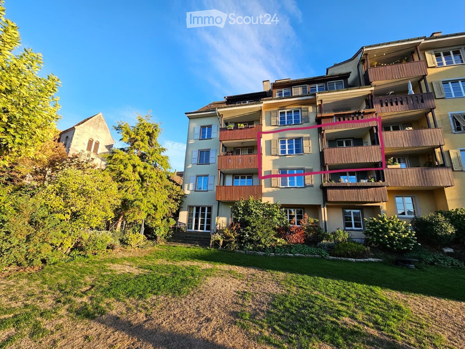 Multi-story residential building with multiple balconies, surrounded by greenery and a lawn.