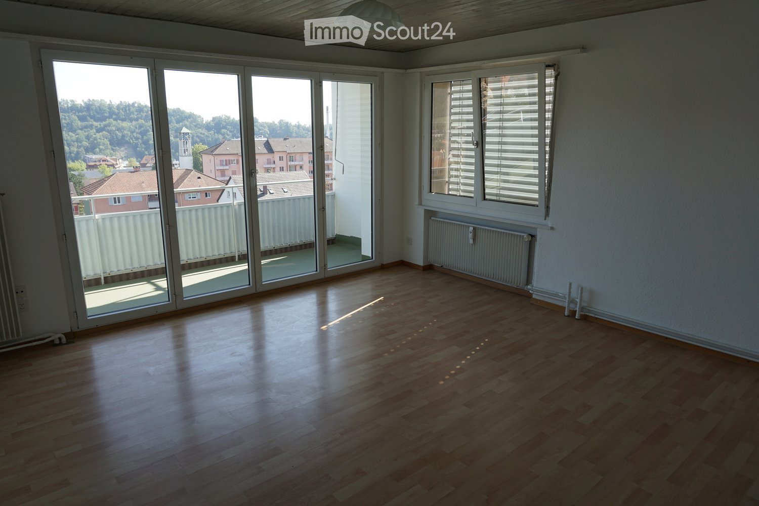 Empty room with wooden flooring, large sliding glass doors leading to a balcony, and a radiator on the wall.