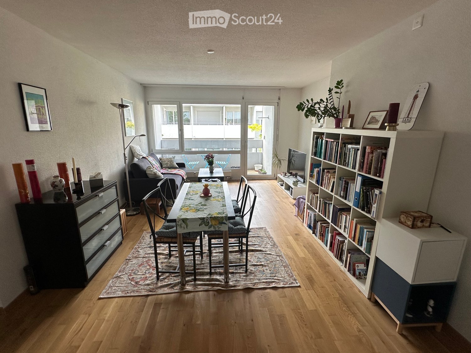 Bright, open living and dining area with wooden flooring, large windows, and a balcony access. Features a sofa, dining table with chairs, bookshelf, cabinet, floor lamp, and television.