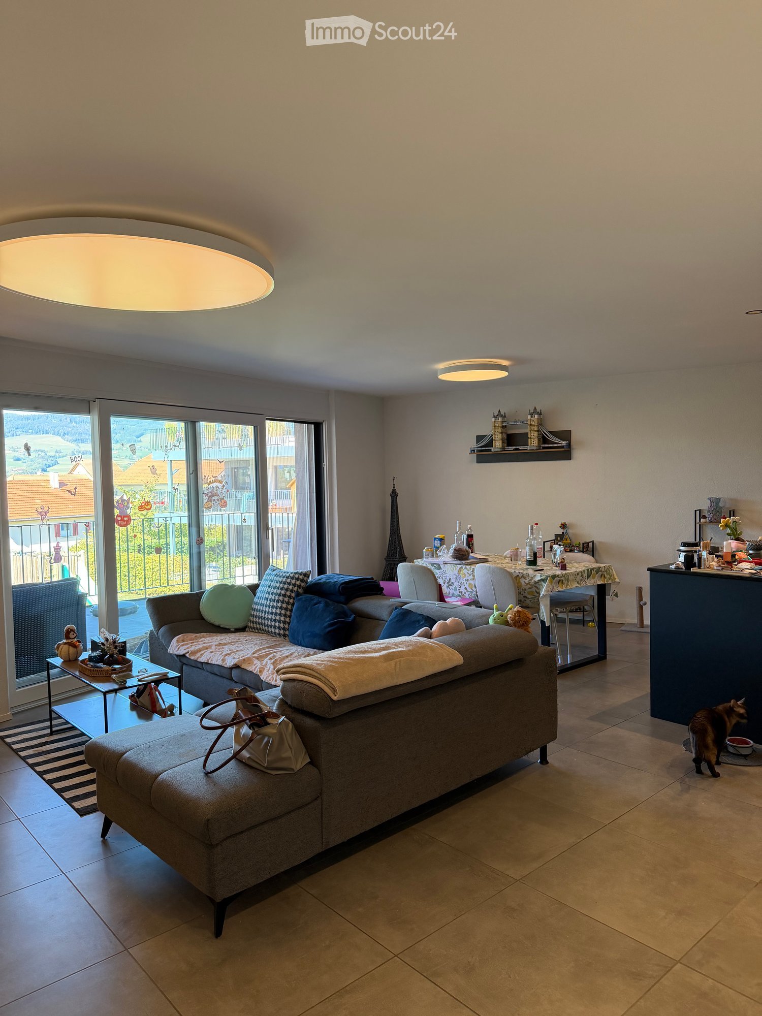 Modern living room with a grey sectional sofa, coffee table, dining area with a table and chairs, and a view of the balcony and outdoor area.