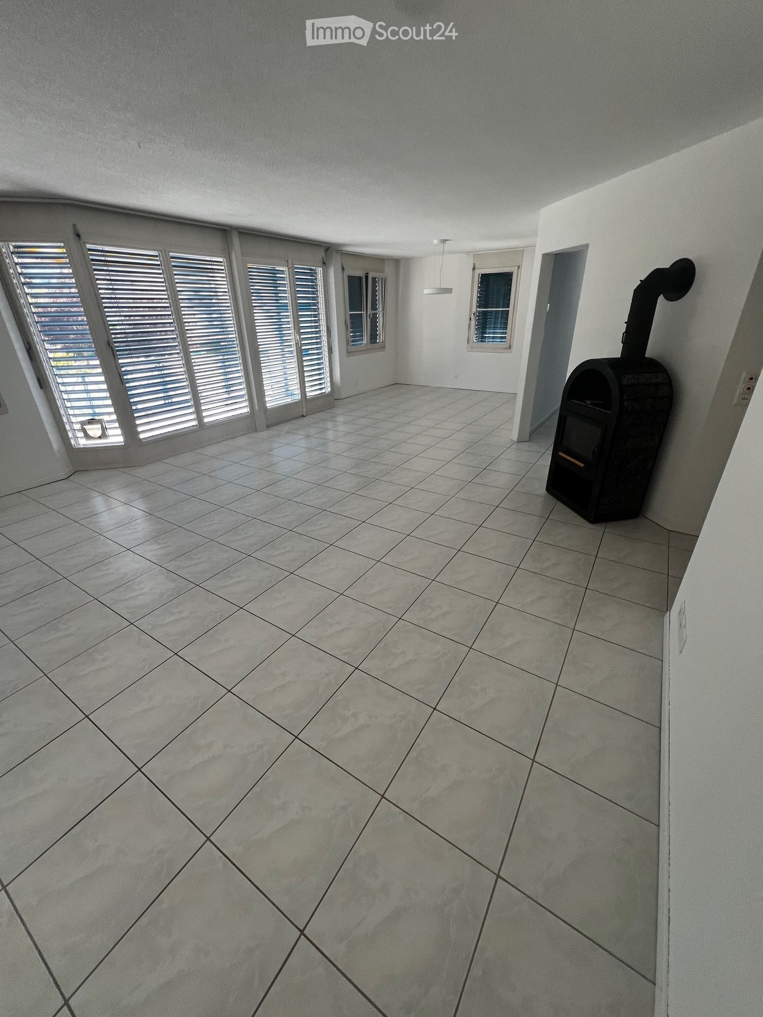 Empty room with tiled floor, large windows with blinds, and a small black stove in the corner.
