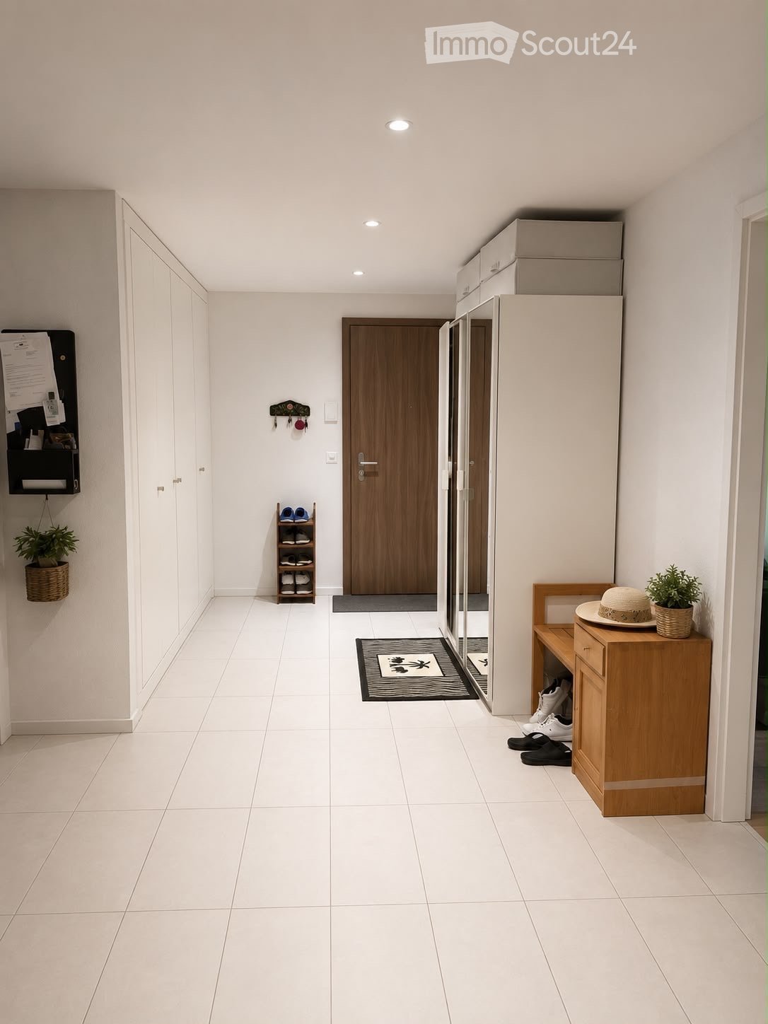 Clean, modern hallway with white tiled flooring, white walls, and recessed ceiling lighting. Features include a wooden door, a small shoe rack, a bench with a hat and plant, and a coat rack with a potted plant.