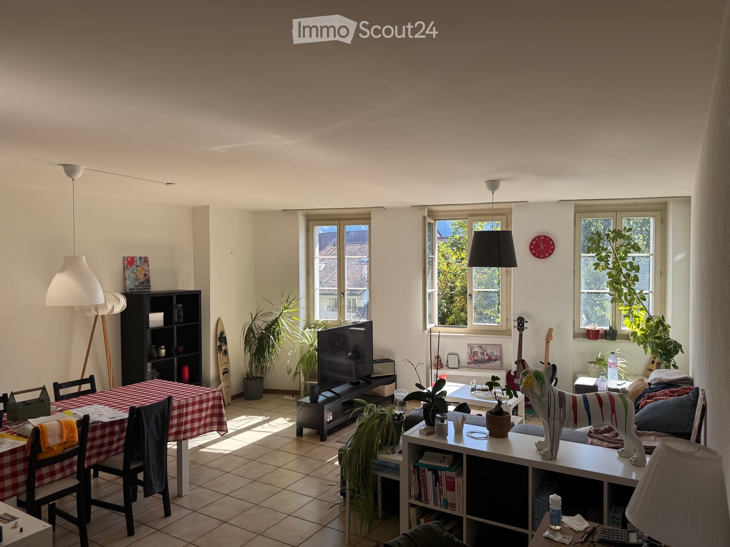 Bright living and dining area with large windows, tiled floor, and modern furniture including a sofa, dining table, TV, bookshelf, and guitars.