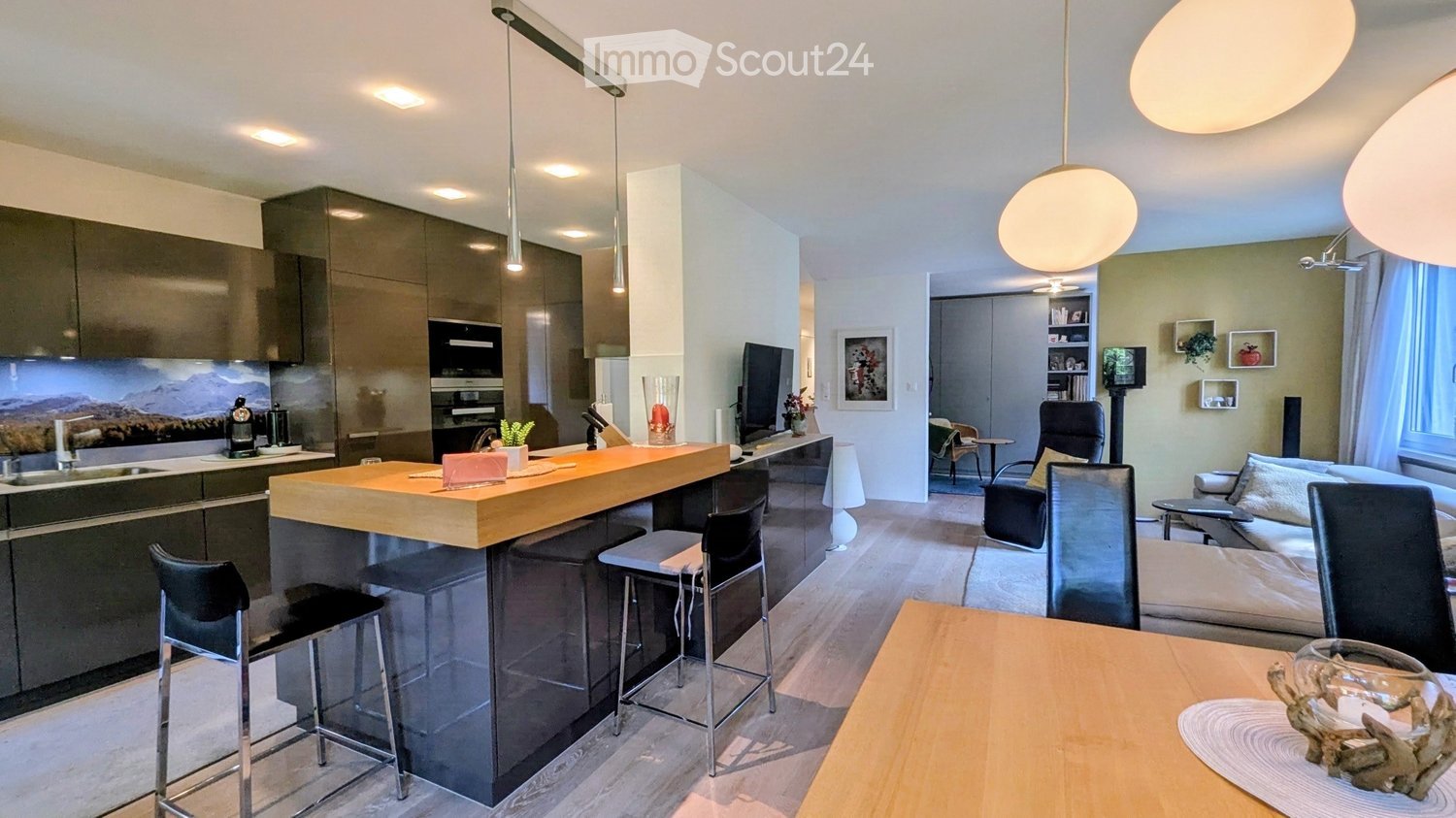 Modern open-plan living and dining area with a kitchen island, bar stools, sofa, coffee table, and dining table. The kitchen features dark cabinets, a sink, and an oven. The living area includes a sofa, a coffee table, and a TV screen.