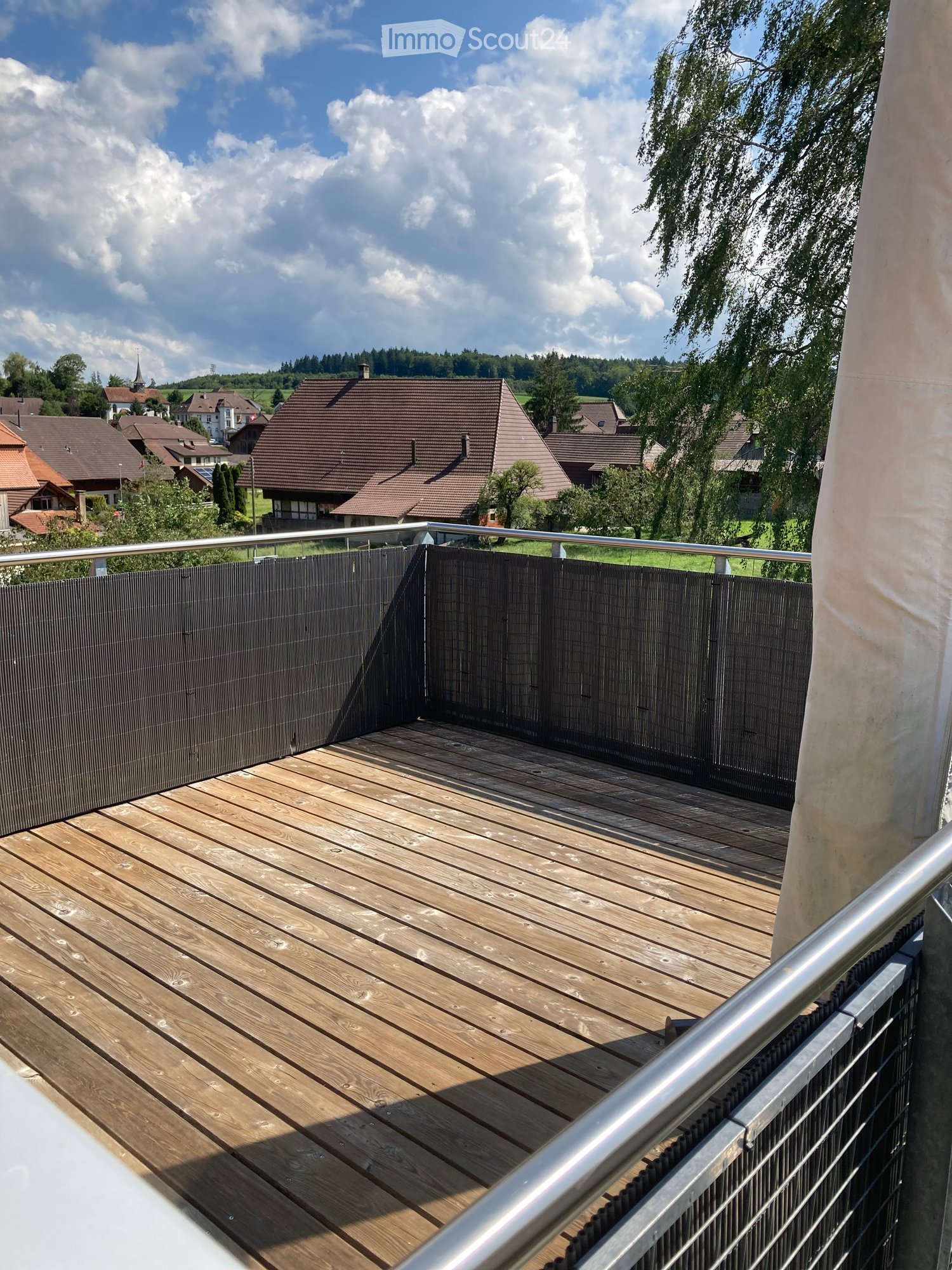 Wooden deck with metal railing, overlooking a residential area with houses and trees under a partly cloudy sky.