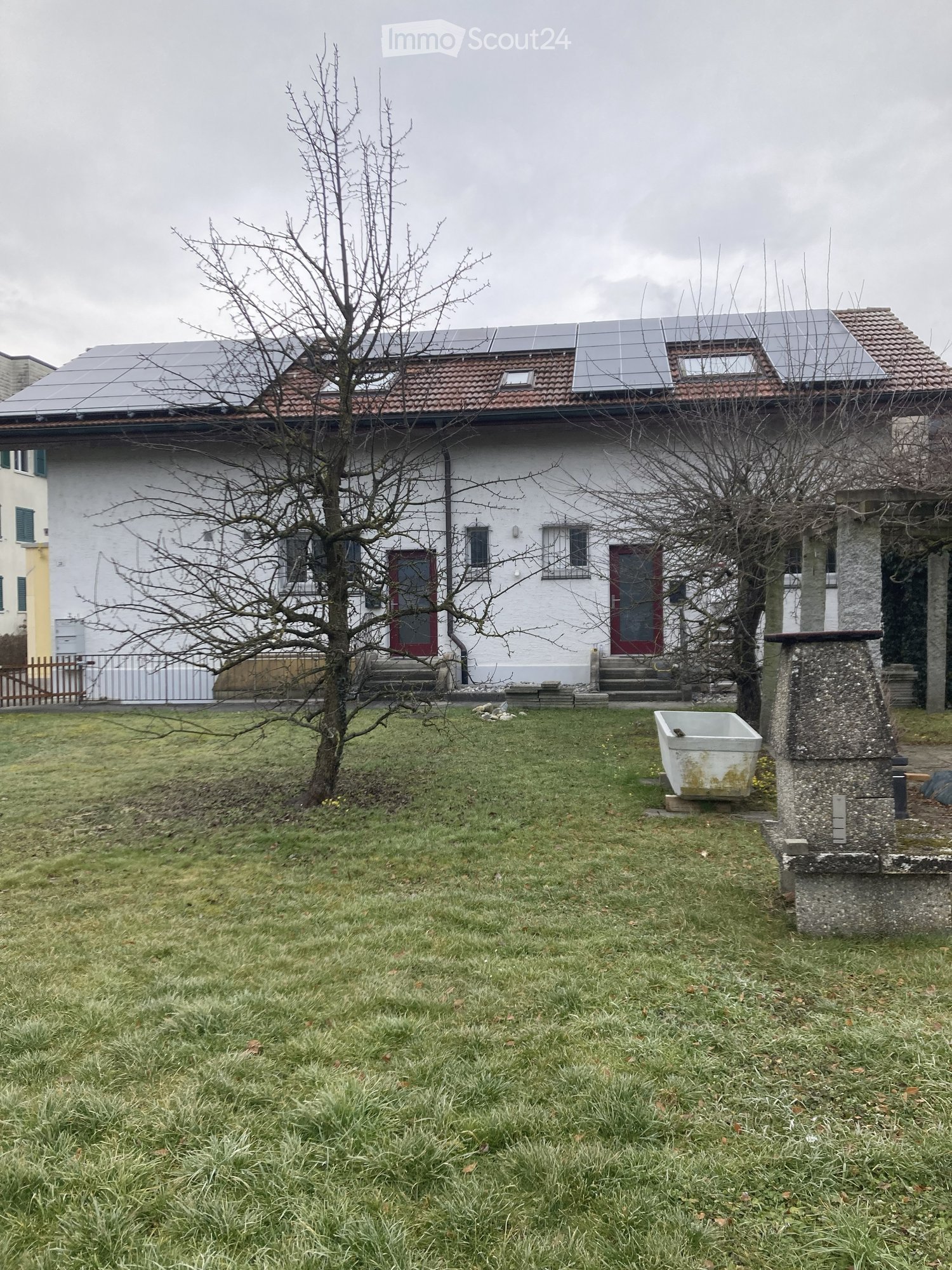Lawn with two leafless trees, a white house with solar panels on the roof, and a small stone structure with a white basin.