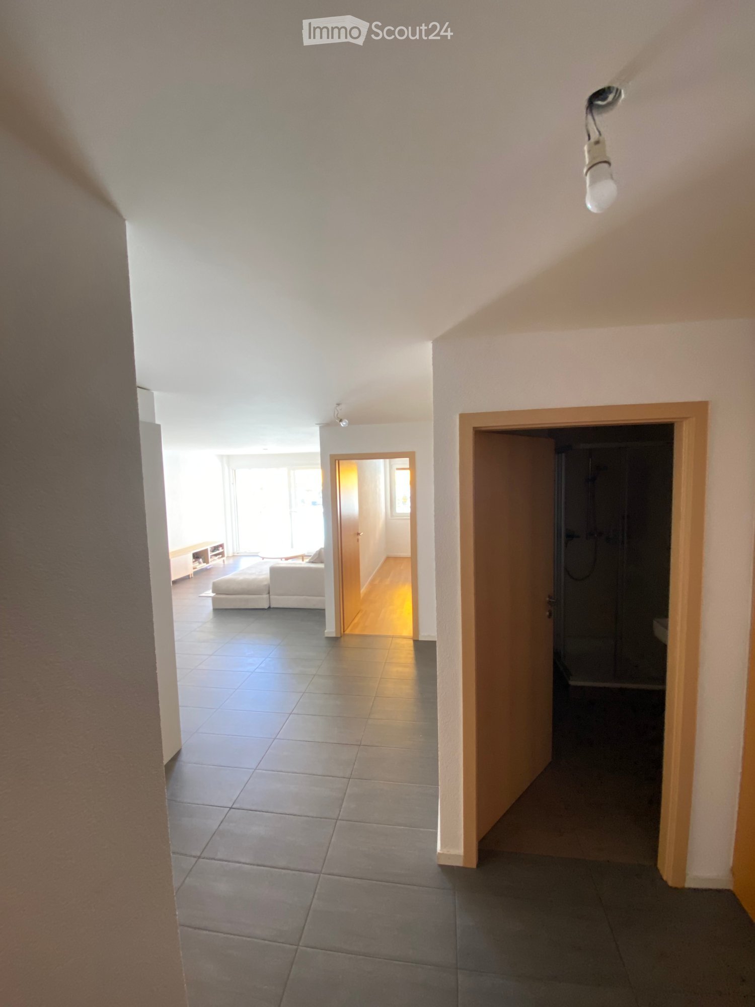 Bright hallway with tiled floor, leading to a living area with white seating and a small table, and a bathroom with a shower stall.
