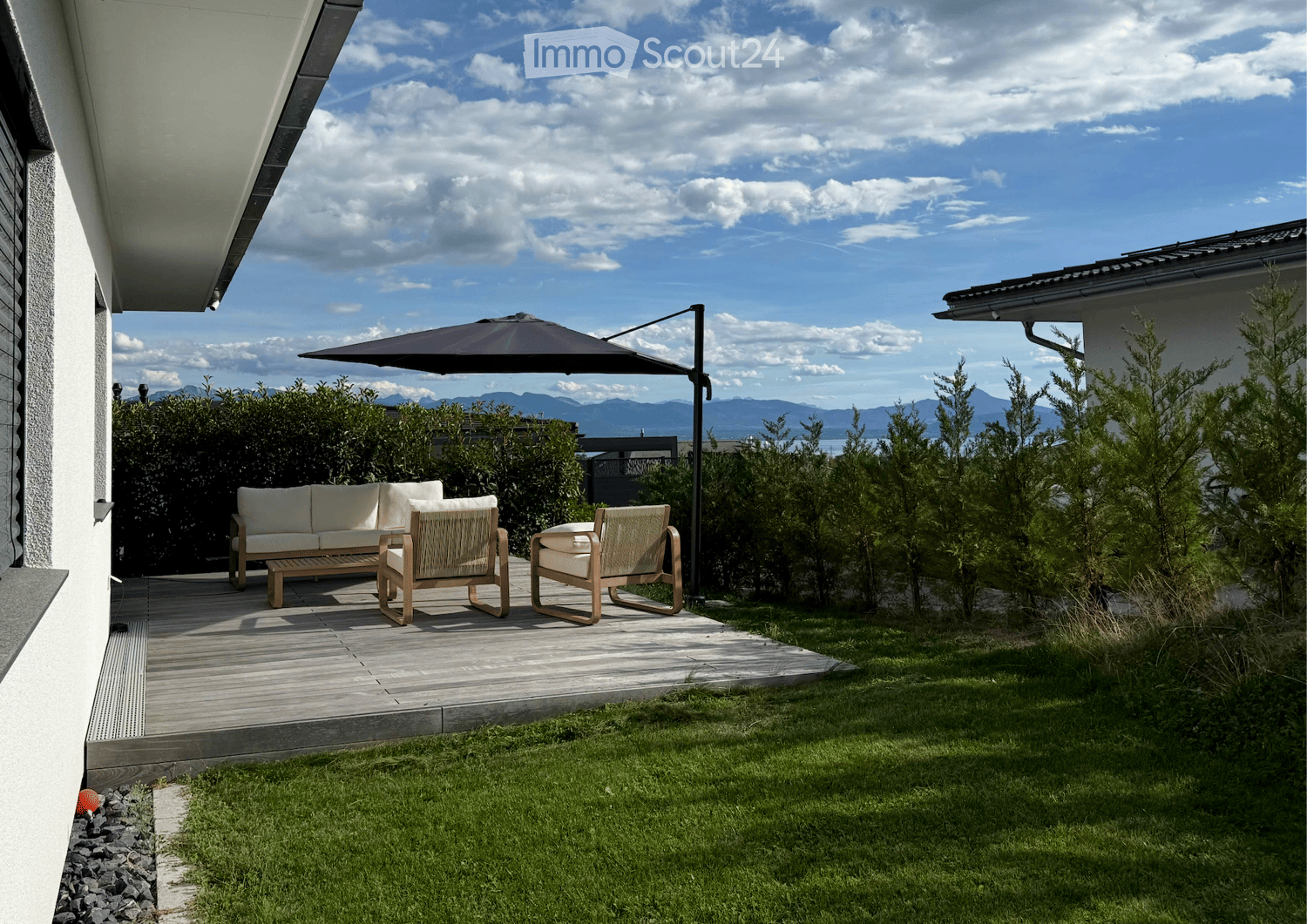 A modern outdoor patio with a wooden deck, white sofa, two wicker chairs, and a large umbrella, surrounded by greenery and a scenic mountain view.