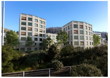 Modern multi-story residential building with white facade, large windows, and surrounding greenery.