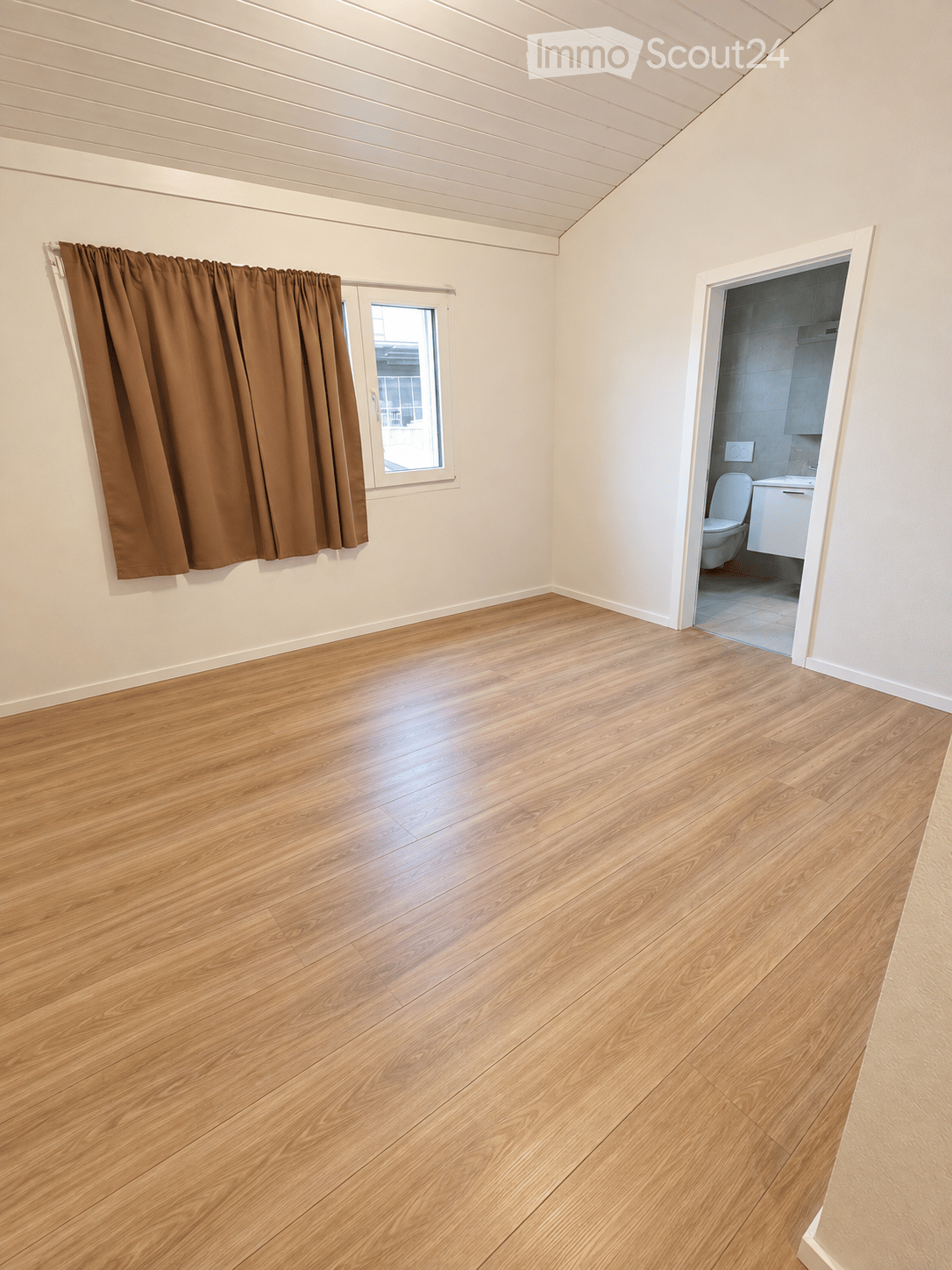 Empty room with wooden flooring, white walls, and a window with brown curtains. An adjacent bathroom is visible through an open doorway.