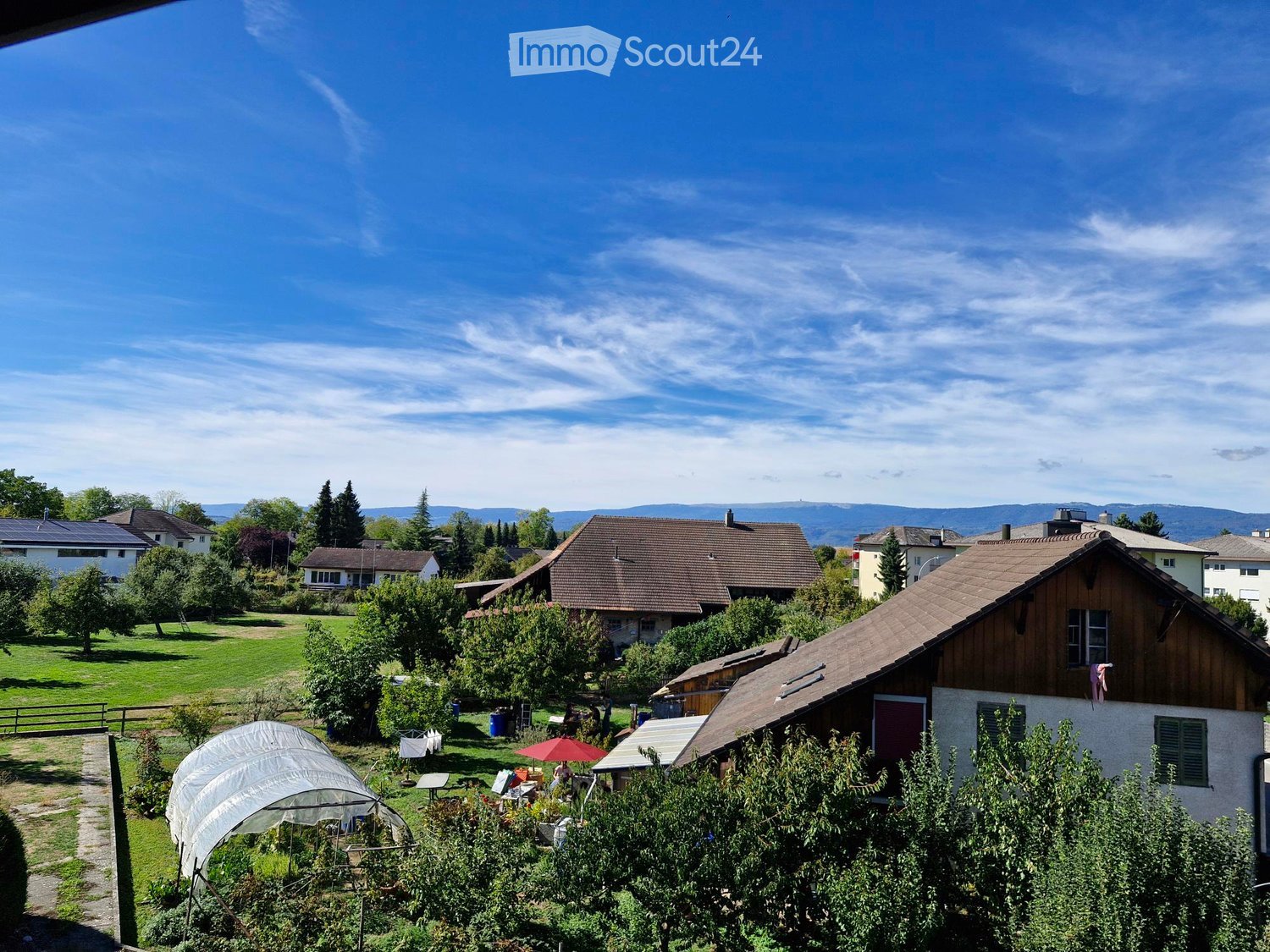 A peaceful garden area with a greenhouse, outdoor furniture, and surrounding houses under a clear blue sky.
