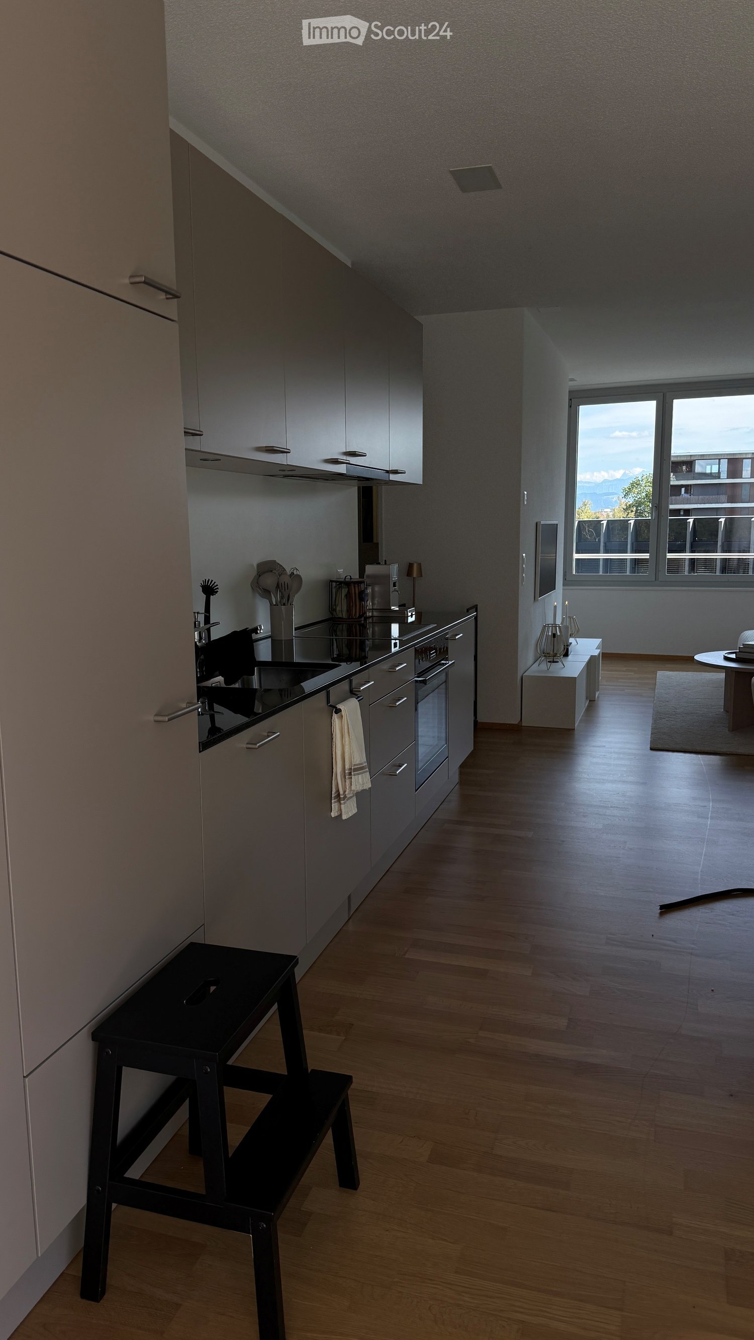 Modern kitchen with grey cabinets, wooden flooring, and a black stool near the sink. A small table and chairs are visible in the adjacent living area.