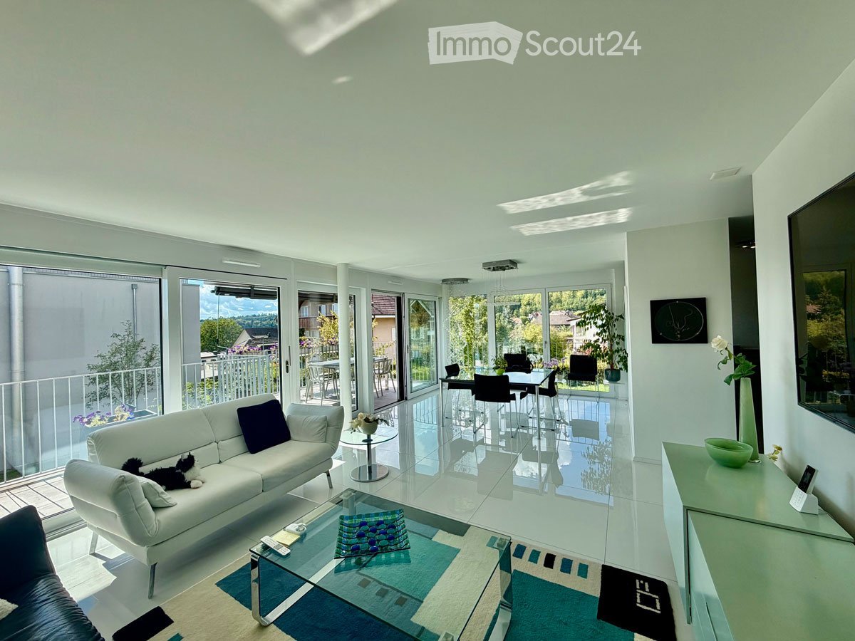 Modern living room with white walls, large windows, and a balcony. Features a white sofa, coffee table, dining area with chairs and table, and a TV screen.