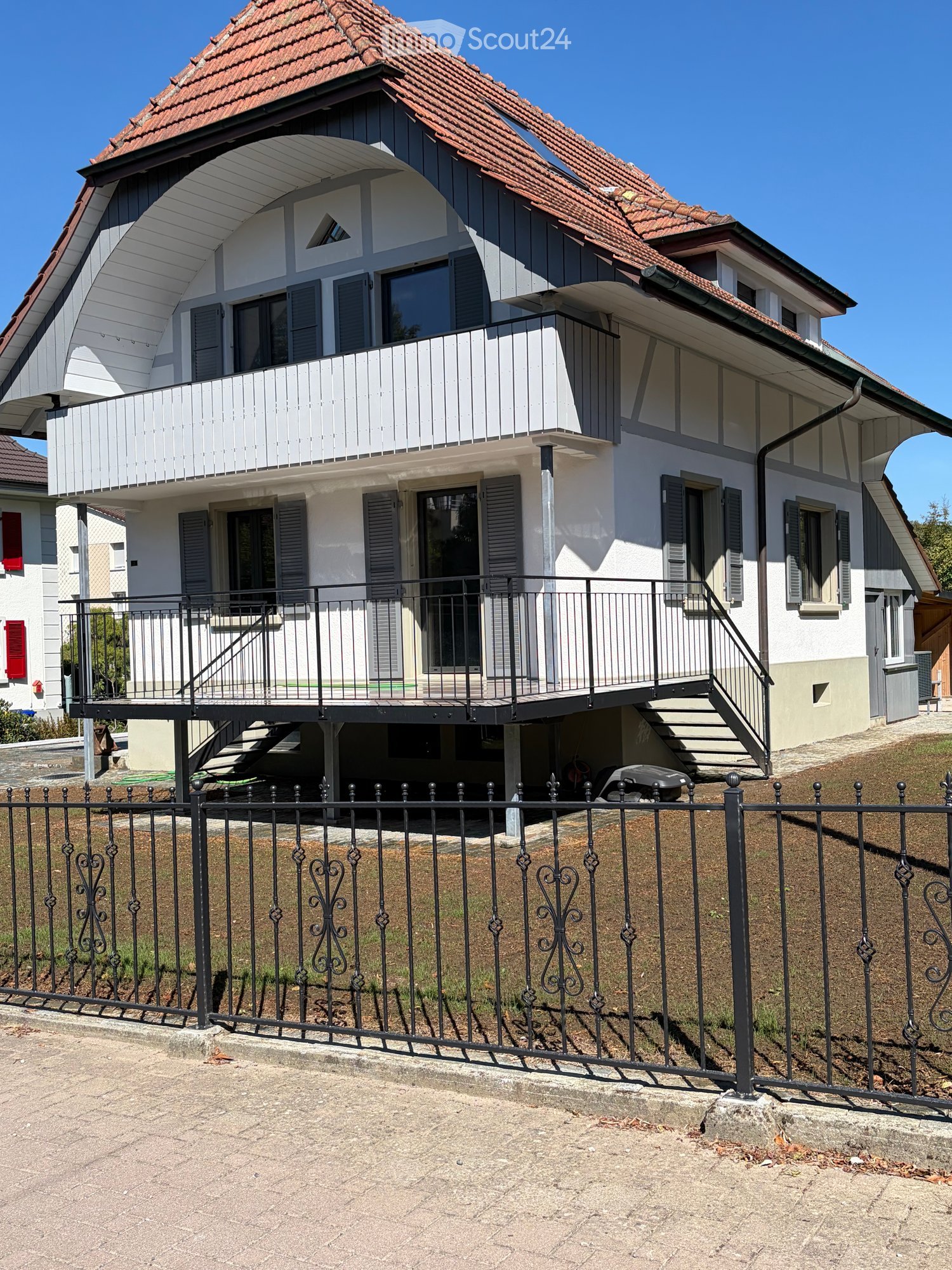 Two-story house with white and grey exterior, red-tiled roof, multiple windows, balconies, and a black metal fence surrounding the property.