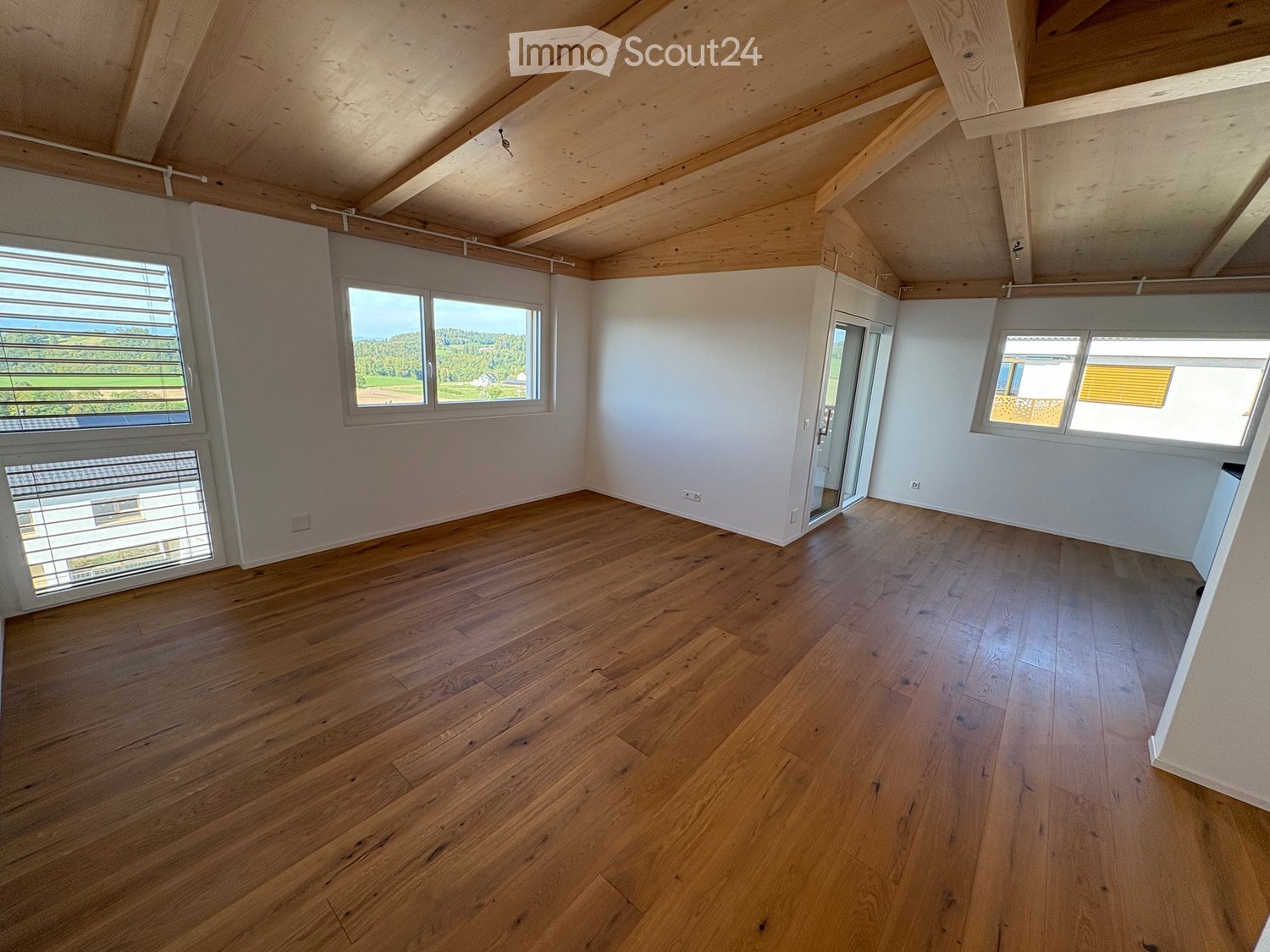 Empty room with wooden flooring, white walls, and large windows offering a scenic view. The ceiling features exposed wooden beams.