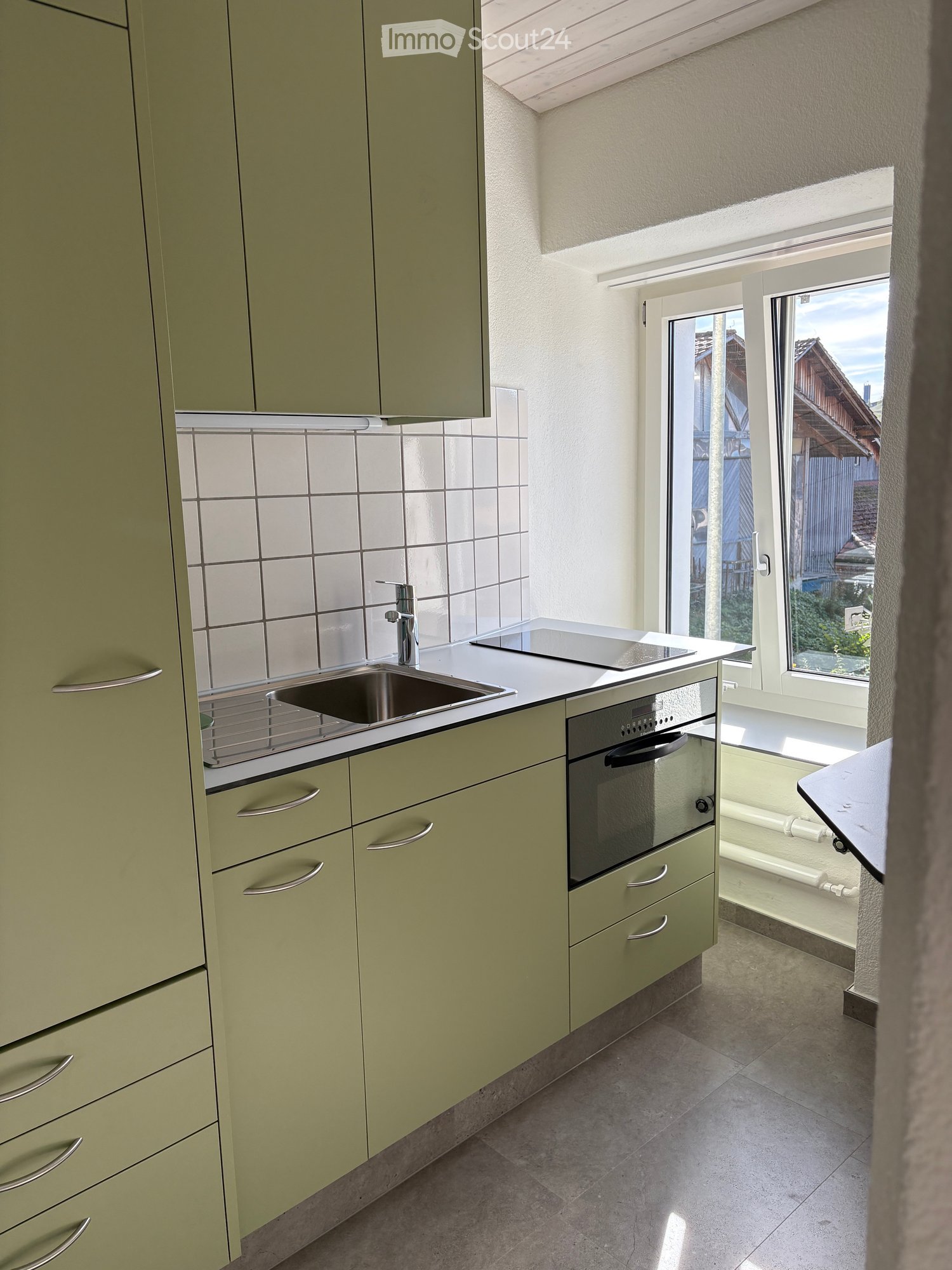 Modern kitchen with light green cabinets, stainless steel sink, oven, and countertop. Features a large window with a view of the outside.