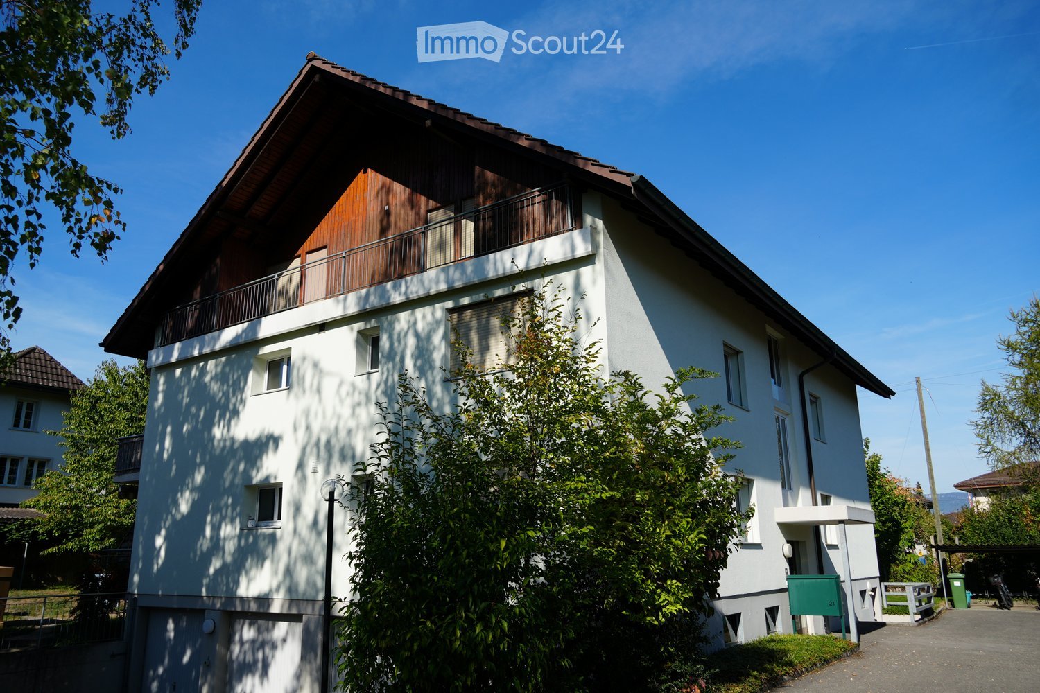 Two-story house with white walls, wooden balcony, multiple windows, and surrounding greenery.