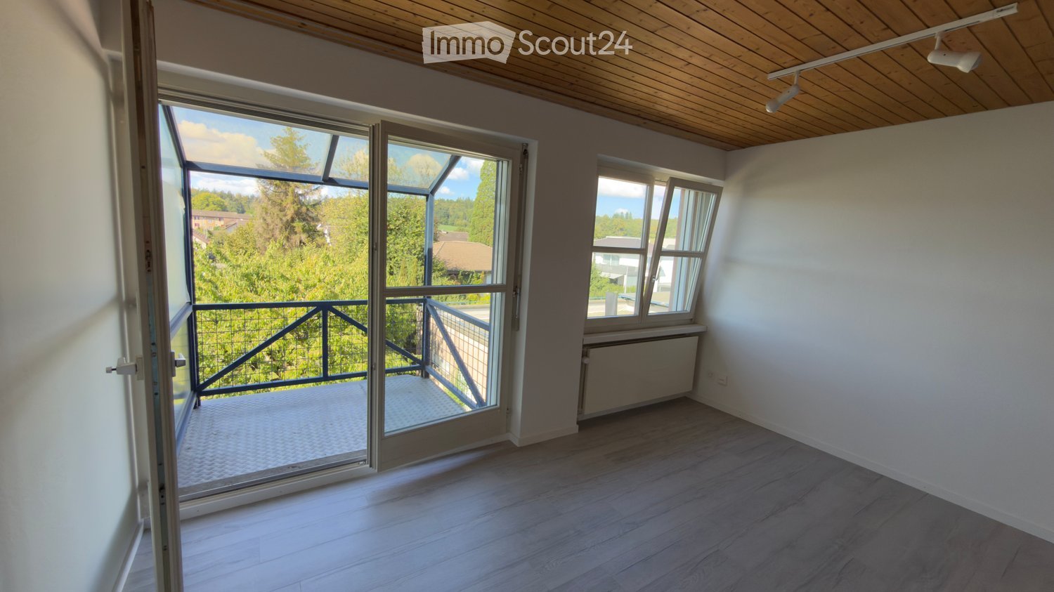 Empty room with wooden flooring, white walls, large windows, and a sliding glass door leading to a balcony with a metal railing.