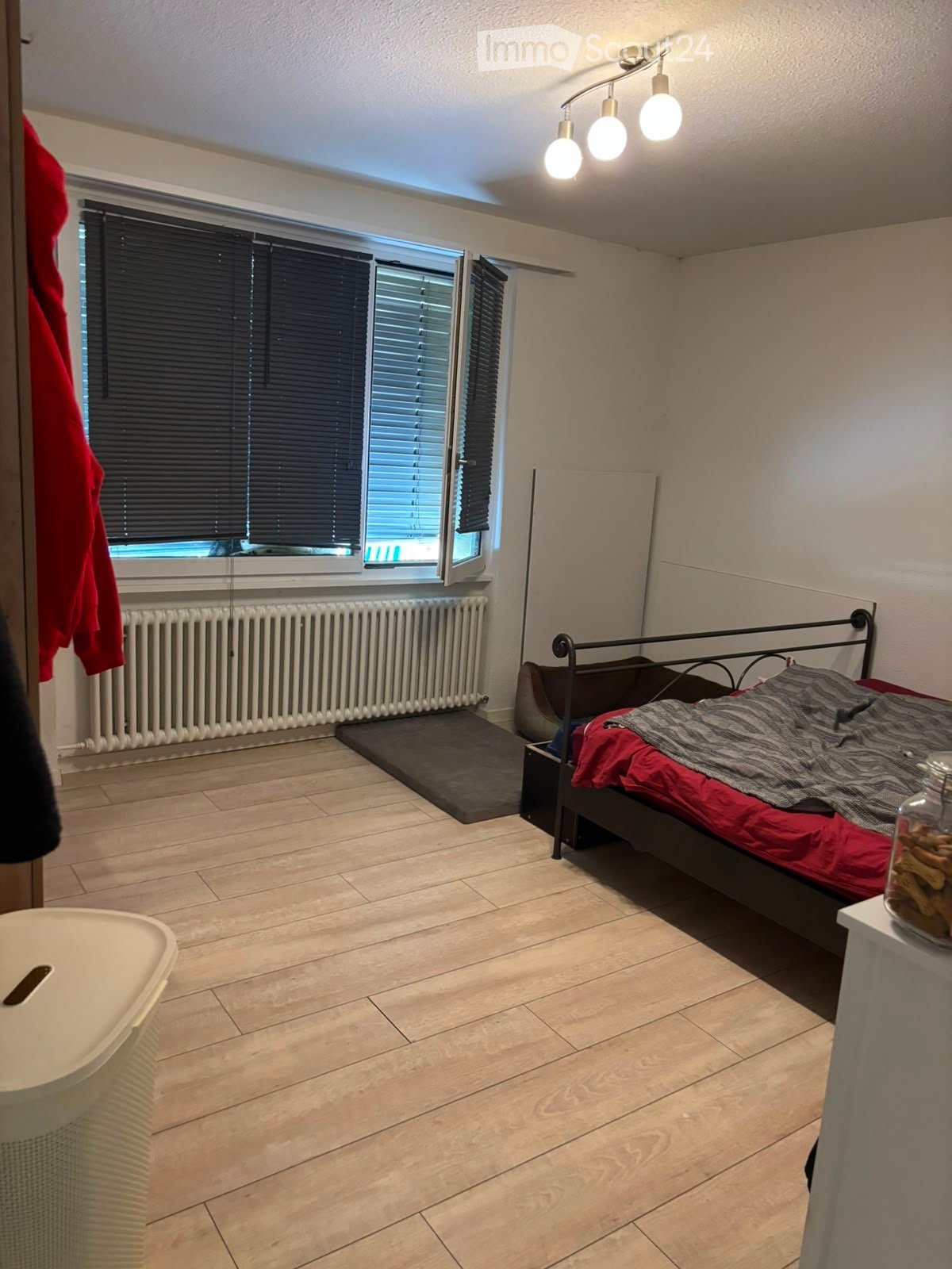 Bedroom with tiled floor, large window with blinds, radiator, bed with red and grey bedding, nightstand with jar, and a white trash bin.