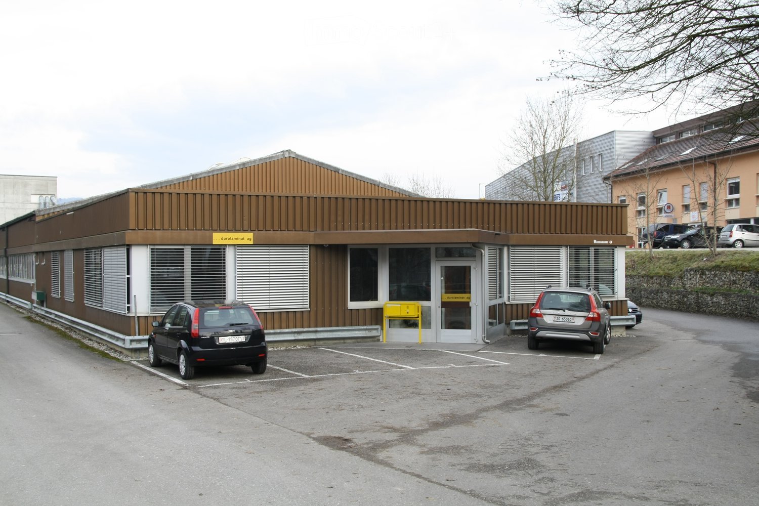 A modern, single-story building with a brown wooden facade and white trim, featuring large windows and a central entrance. Two cars are parked in front of the building.