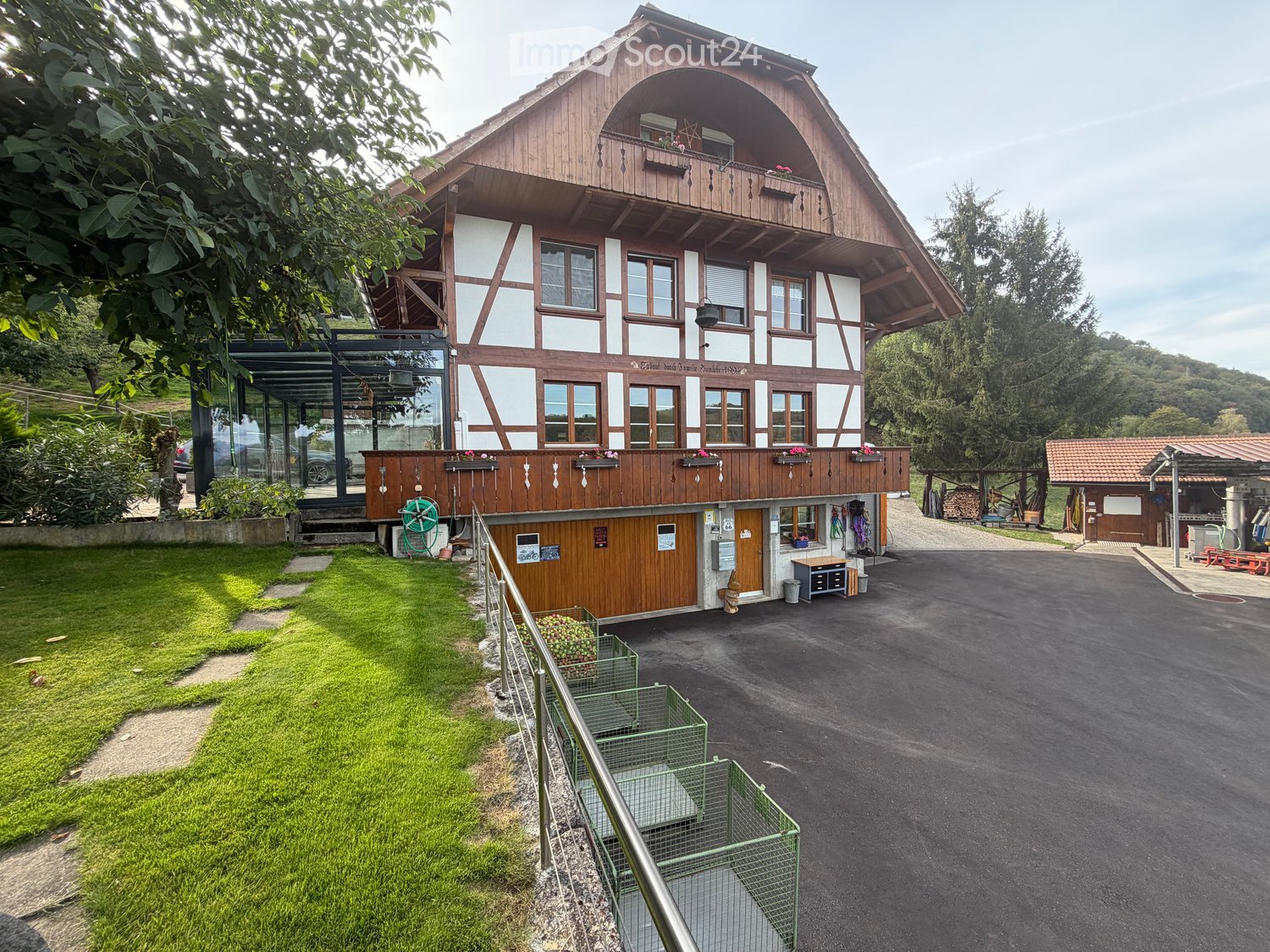 A two-story wooden house with white trim, large windows, and a balcony. The house features a modern glass extension on the left side and is surrounded by a well-maintained lawn and trees.