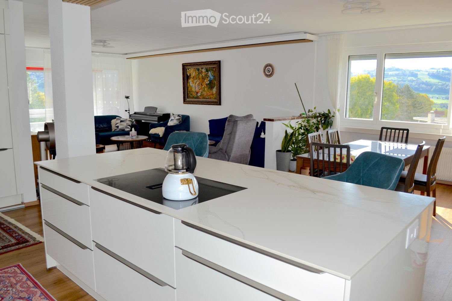 Open-plan living and dining area with a modern kitchen island, sofas, a piano, and large windows offering a scenic view.