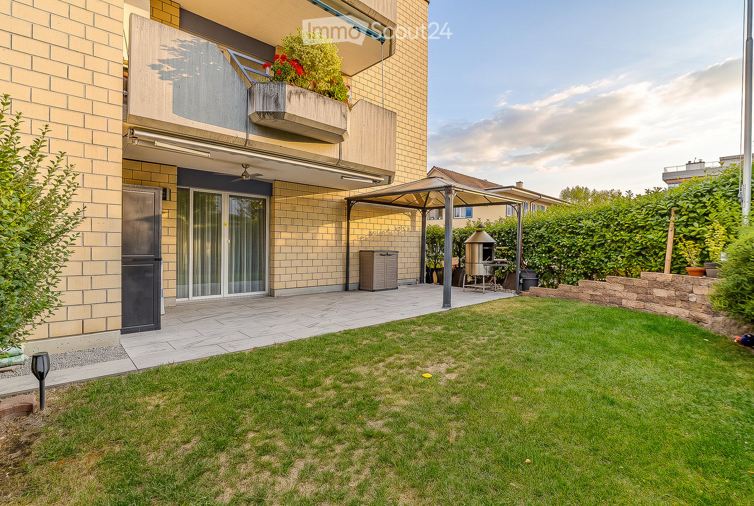 Well-maintained garden with a neatly trimmed lawn, a patio area with a pergola, a barbecue, and a small storage shed. The house features a balcony with plants and flowers, and the exterior is made of light-colored brick.