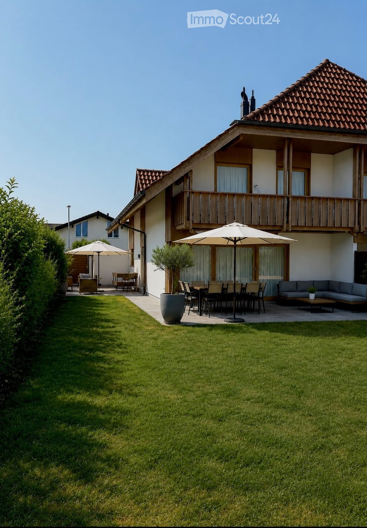 Lush green lawn with patio furniture, including a sofa, dining table, chairs, and umbrellas, near a modern house with a balcony and large windows.