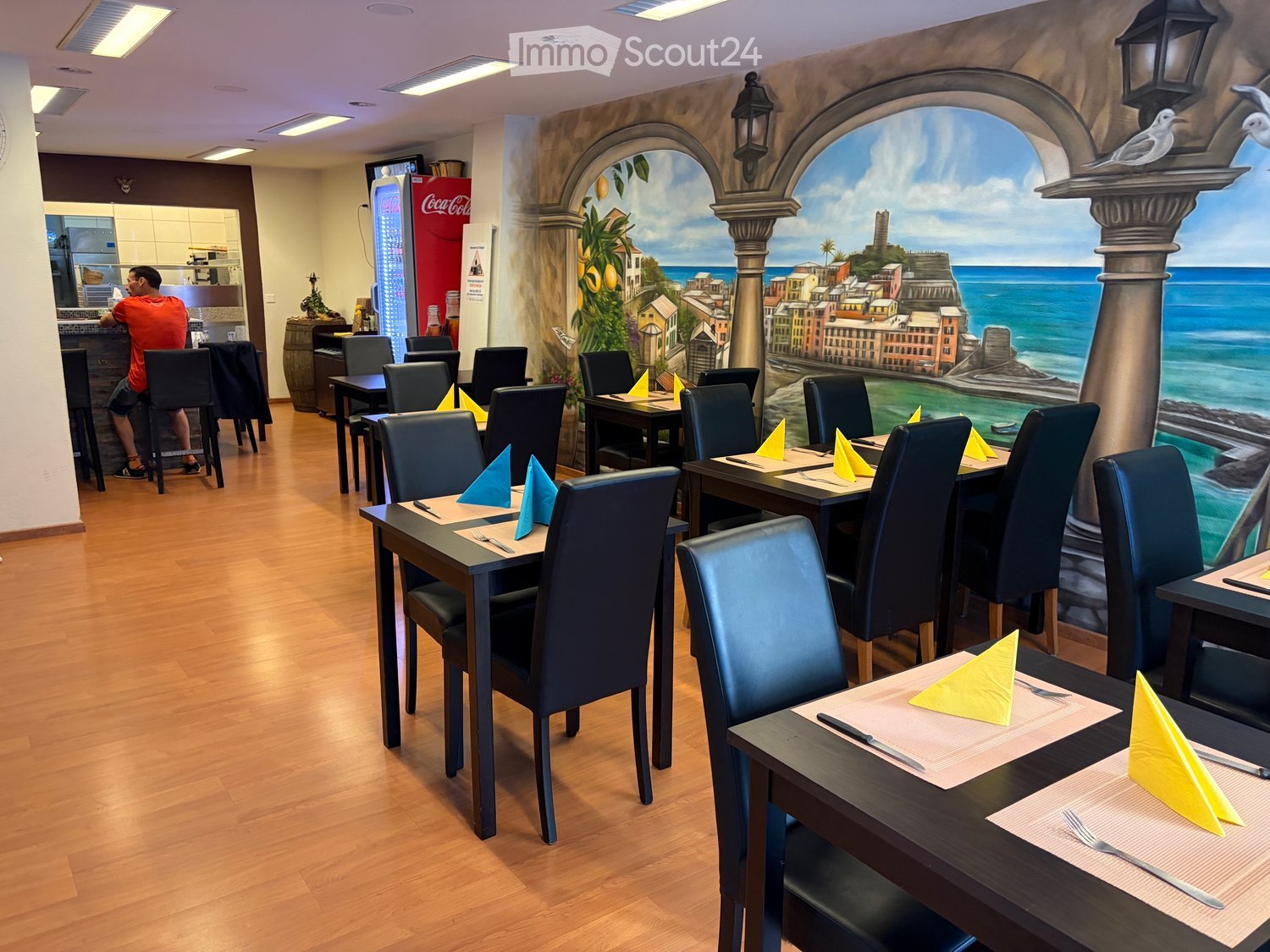 Restaurant interior with wooden floors, tables, chairs, and a mural depicting a coastal scene. Tables are set with napkins, forks, and knives.