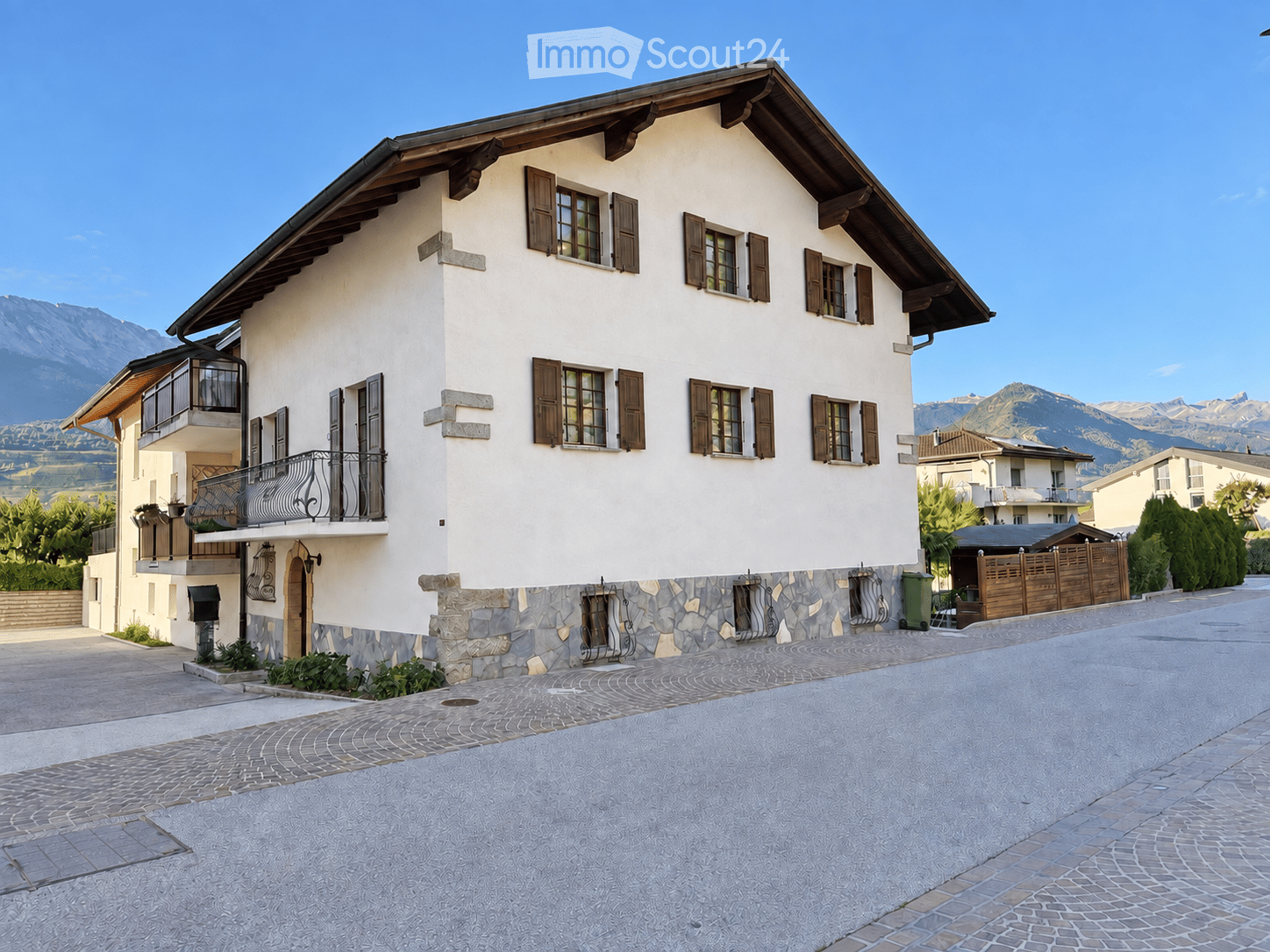 Two-story house with white walls, wooden shutters, stone base, and a balcony overlooking a scenic mountain view.