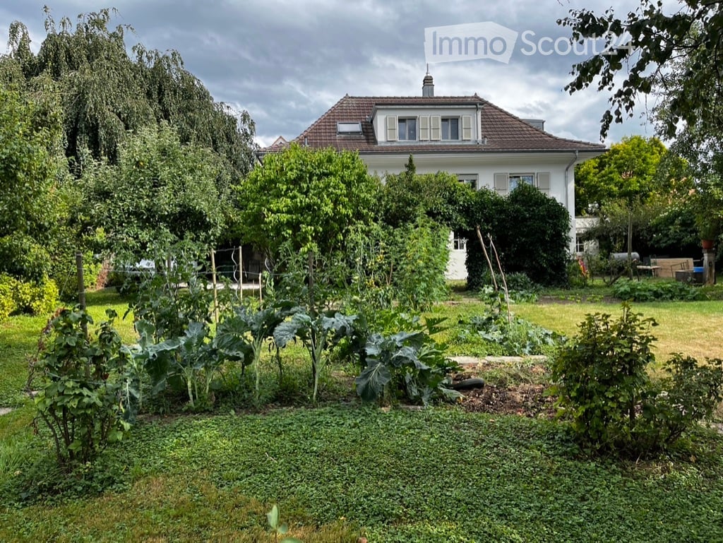 A well-maintained garden with a variety of plants and vegetables, surrounded by a white house with a red-tiled roof and multiple windows.