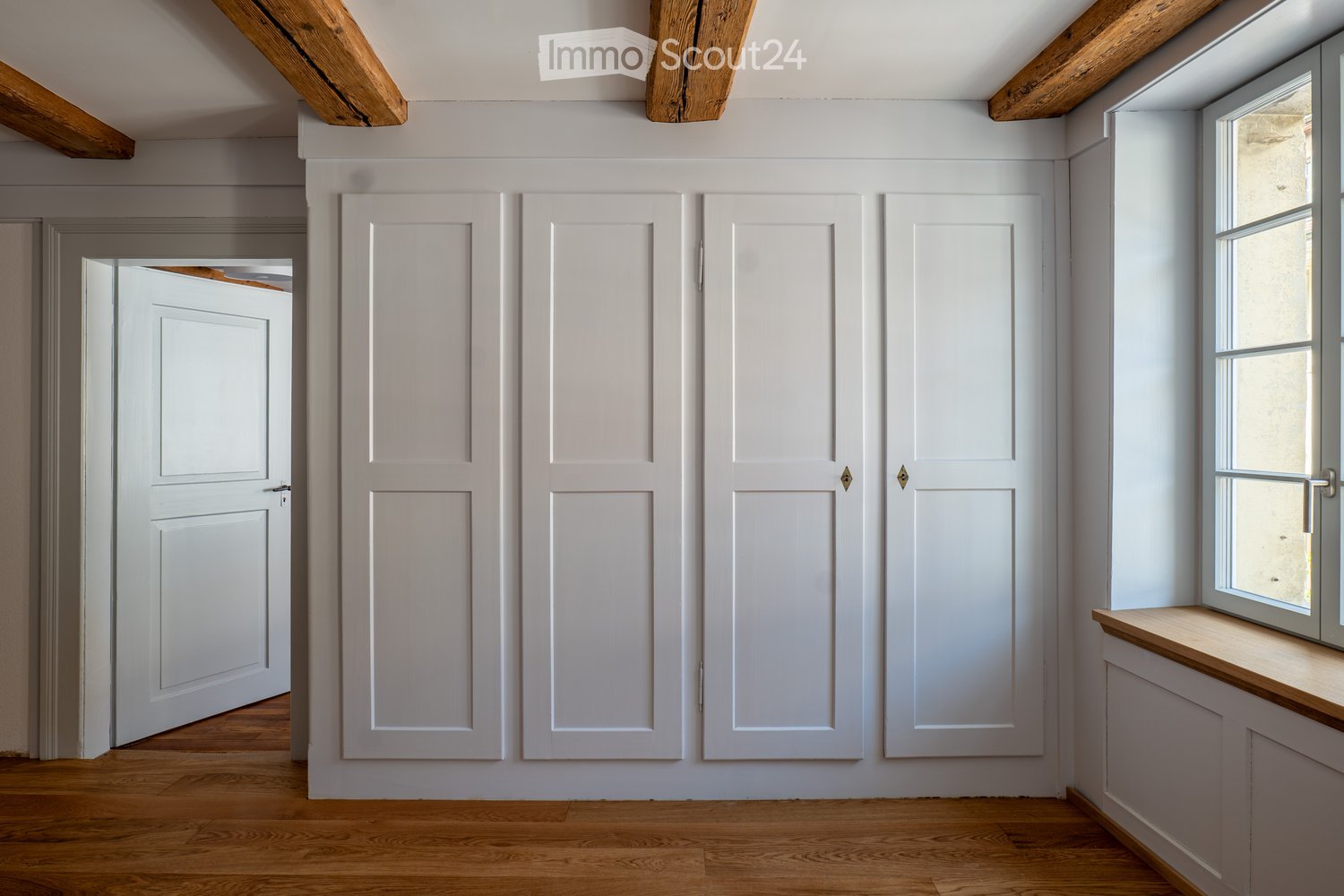 Empty room with wooden flooring, white walls, and exposed wooden beams on the ceiling. Features a window with a wooden sill and a set of white double doors.