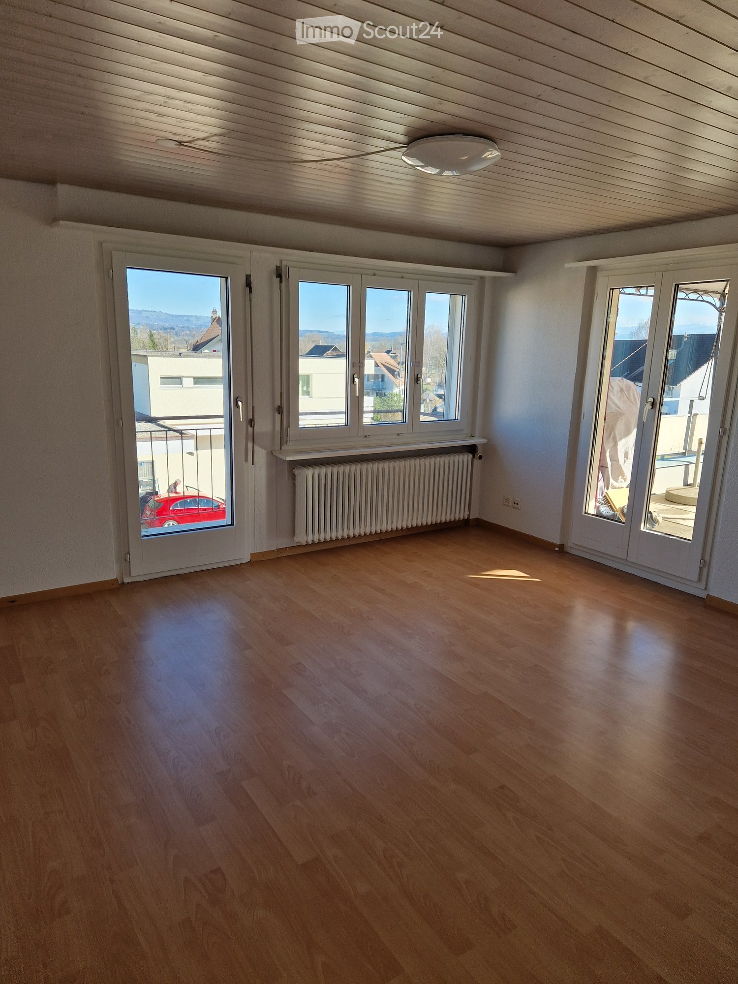 Empty room with wooden flooring, white walls, large windows, and a radiator. The room has a slanted ceiling and a view of the outside through the windows.