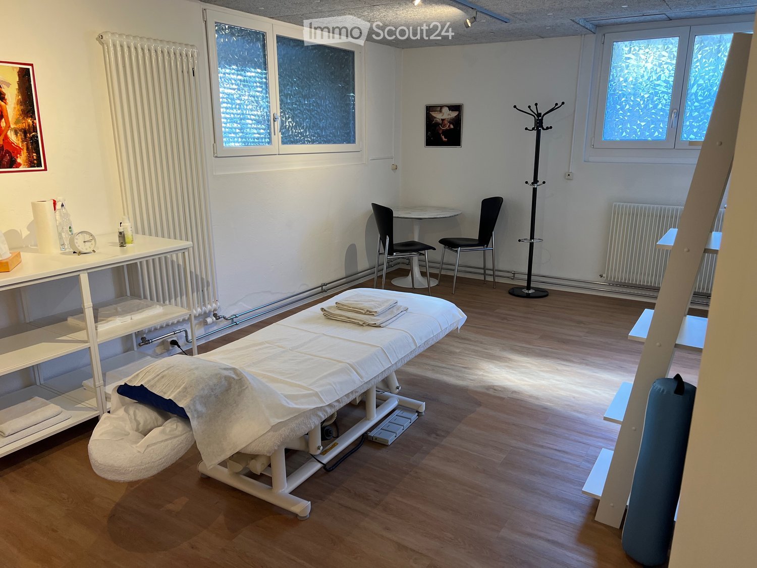 A room with a massage table, two chairs, a small table, shelves, and a radiator.