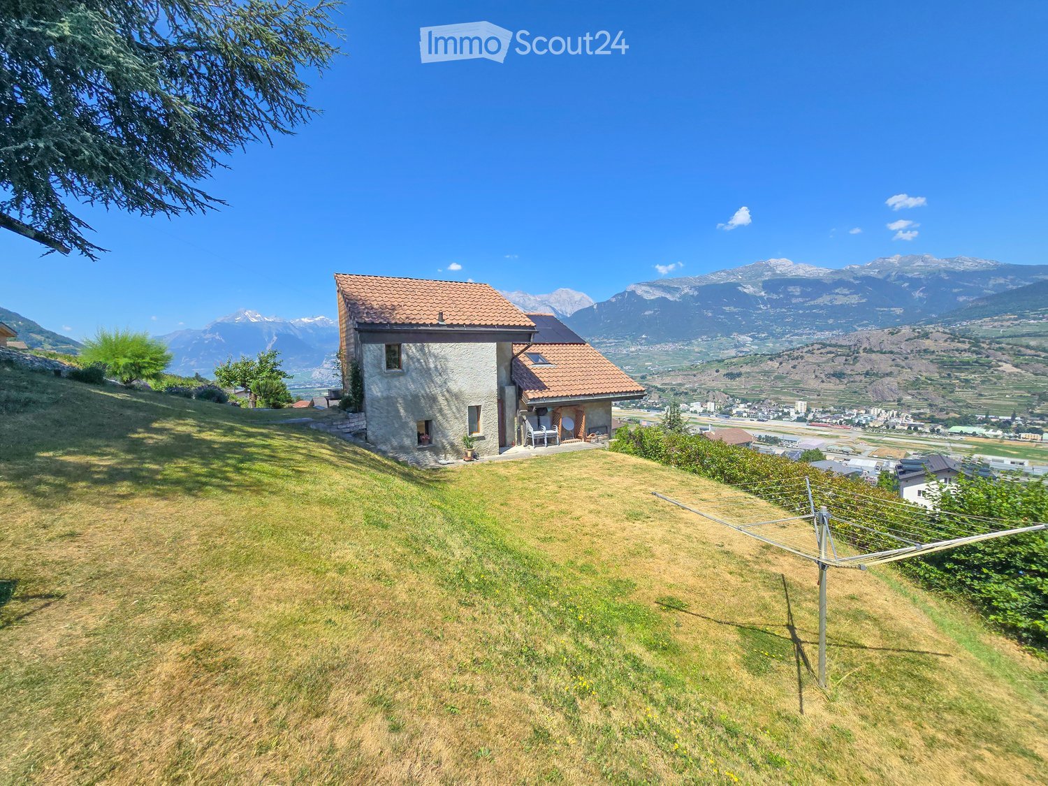 A two-story house with a red-tiled roof and stone walls, situated on a grassy hillside with a panoramic mountain view in the background.